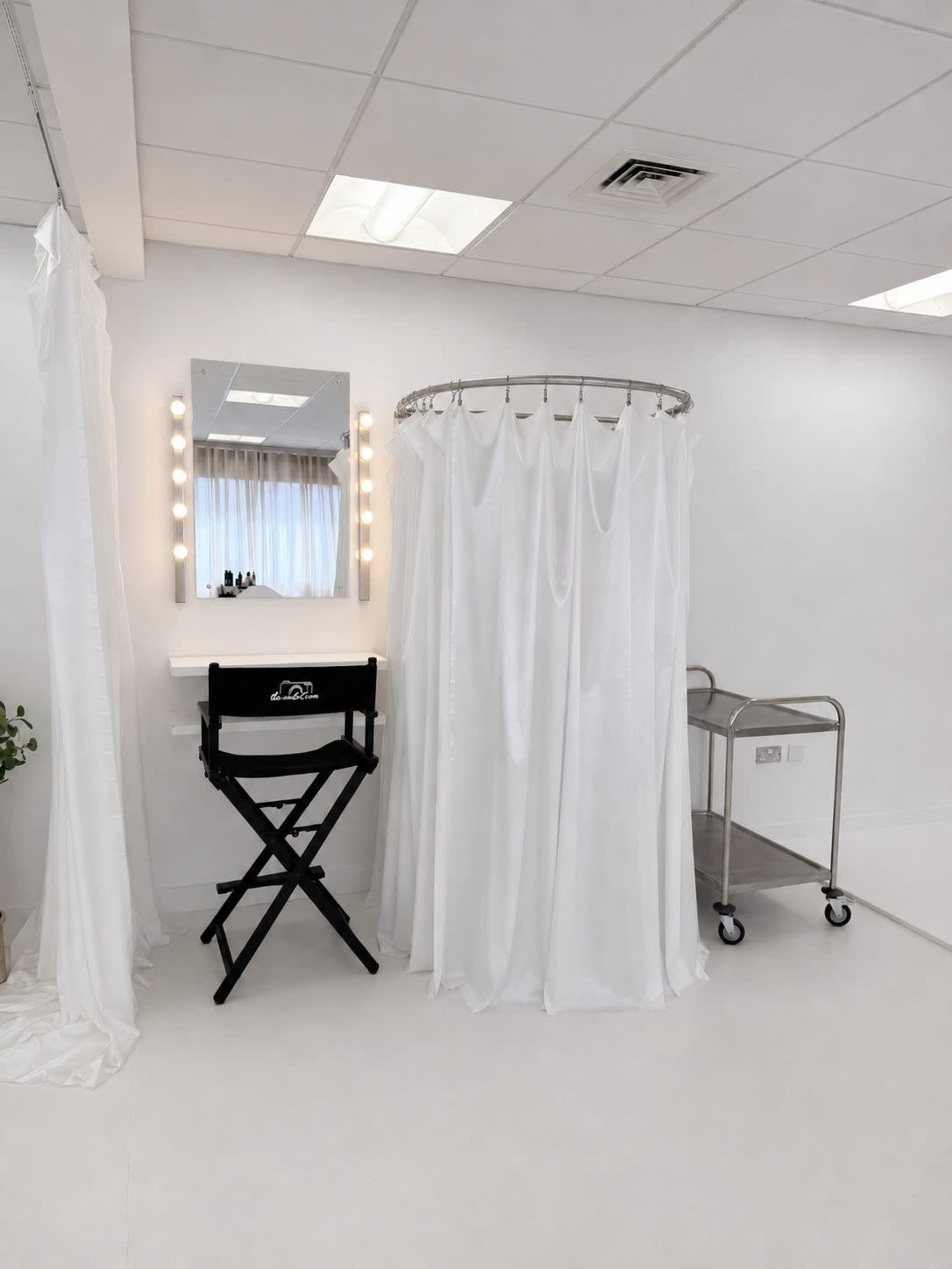 The image shows a minimalist dressing room featuring a round shower curtain, a large mirror with lightbulbs, a black director's chair, and a metal cart.