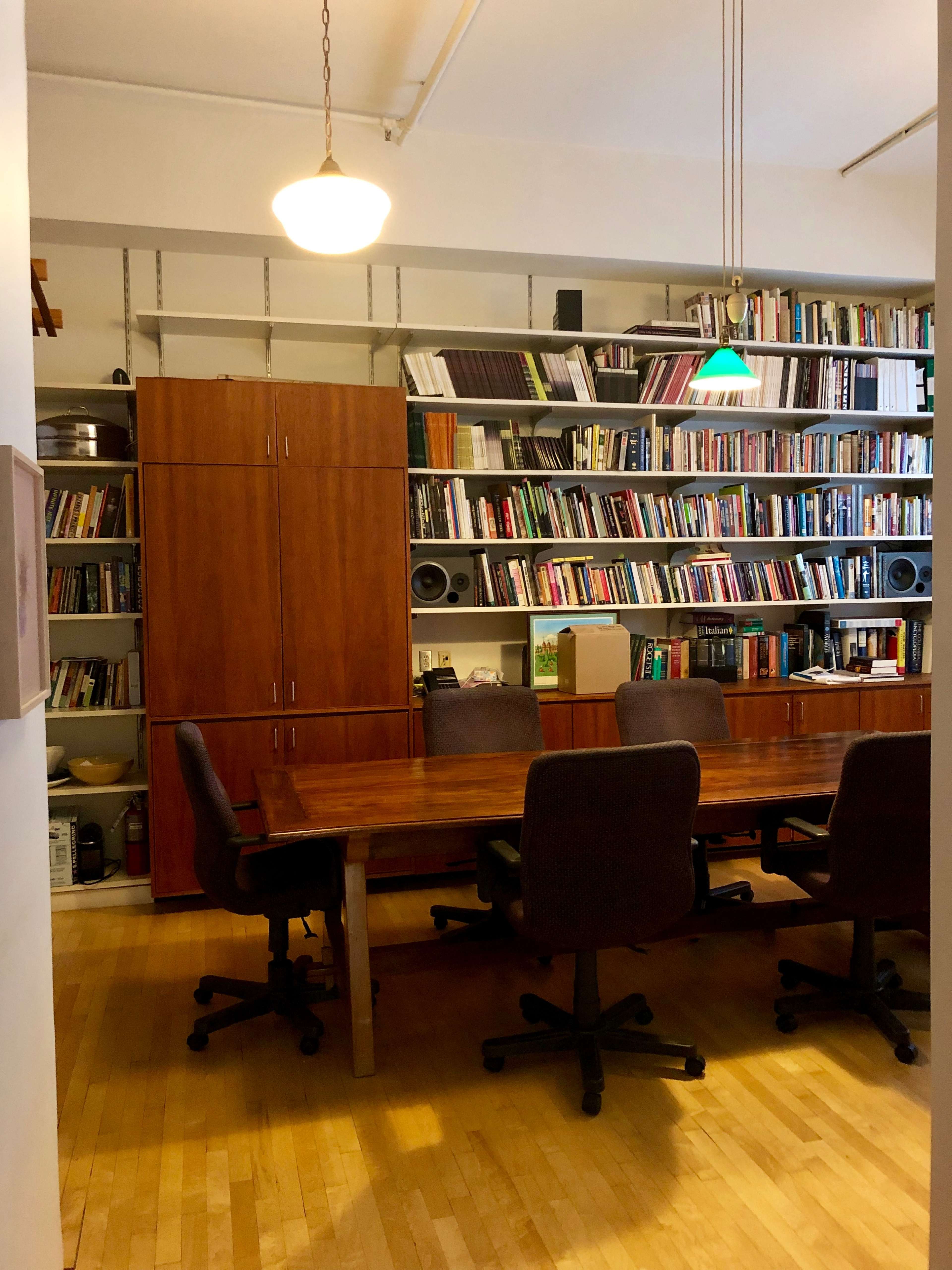A workspace with a large wooden table surrounded by four chairs, facing a wall of bookshelves filled with books and other items.