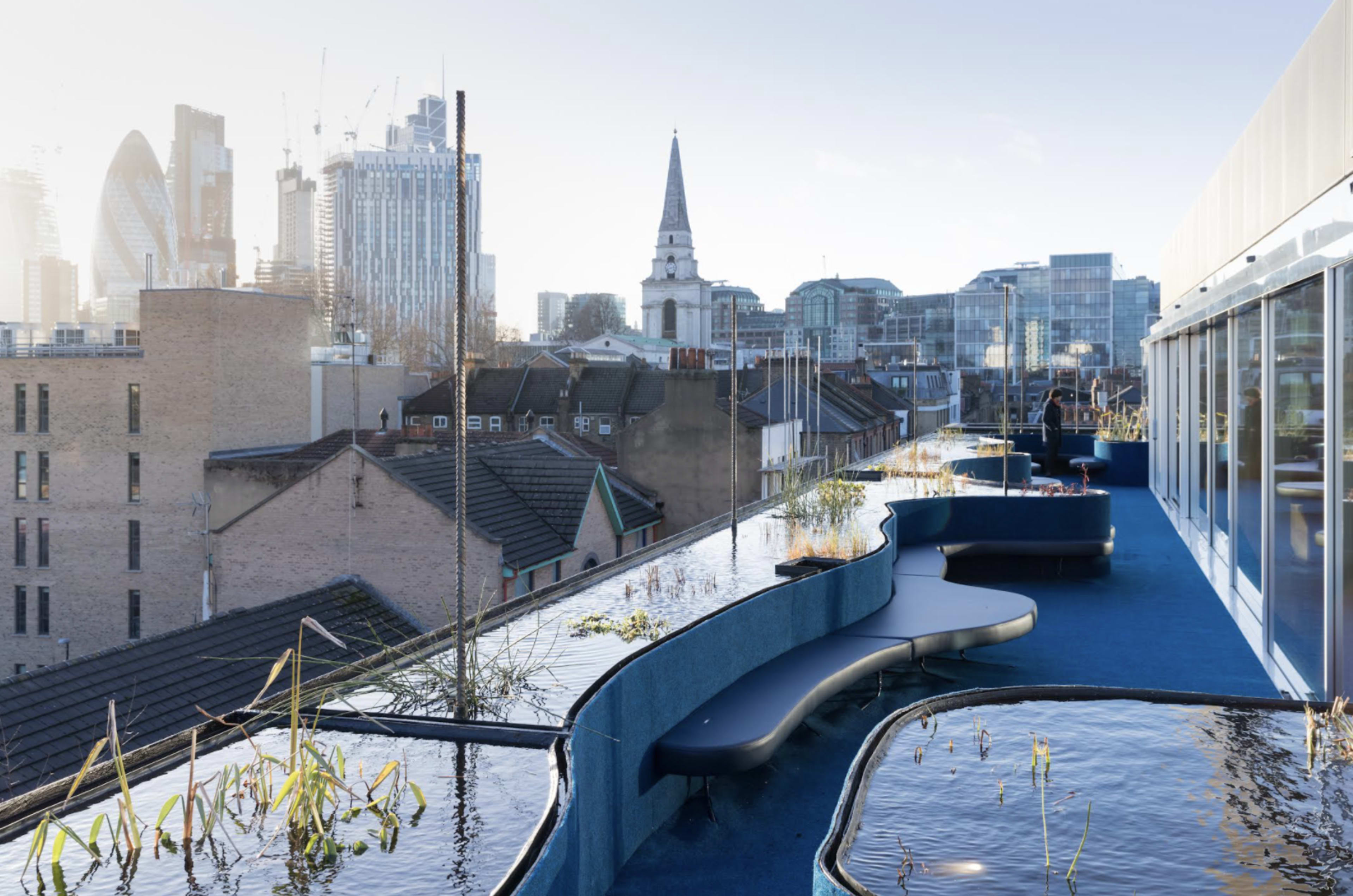 The image shows a rooftop terrace with wavy seating along a water feature, overlooking a city skyline with modern buildings and a church spire.