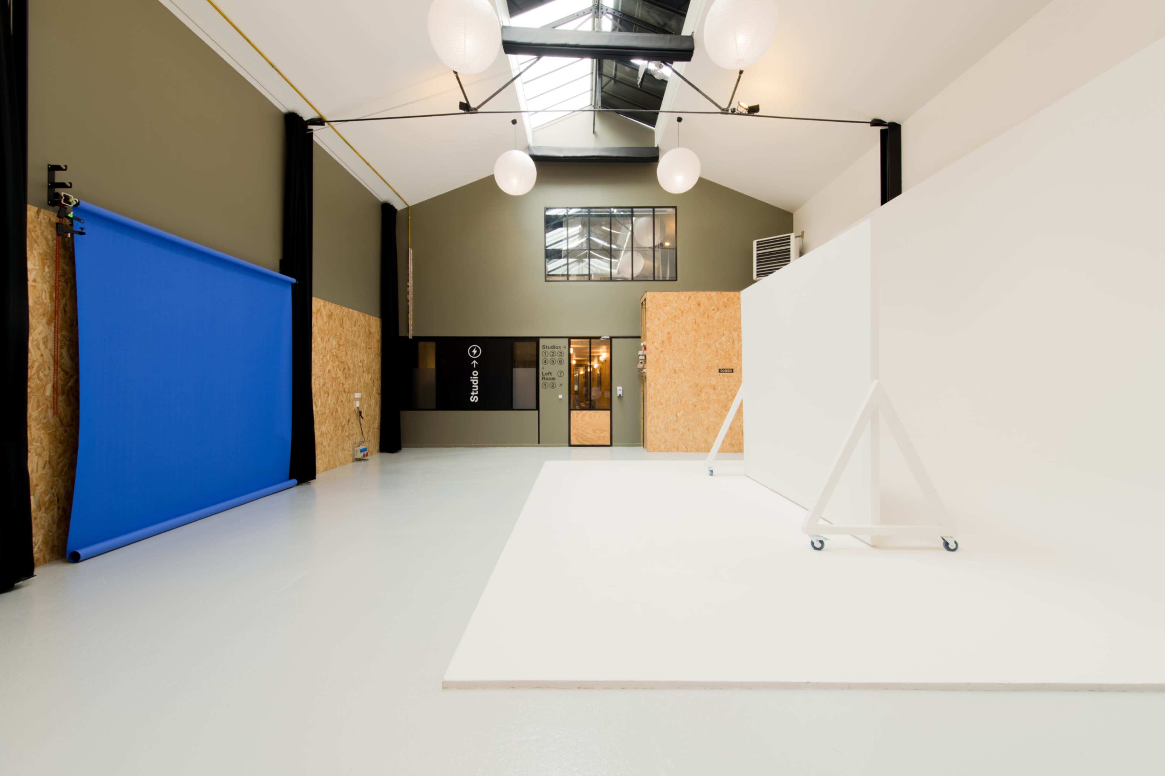 A spacious studio with a blue backdrop on the left, a large white wall, and exposed beams under a skylight.