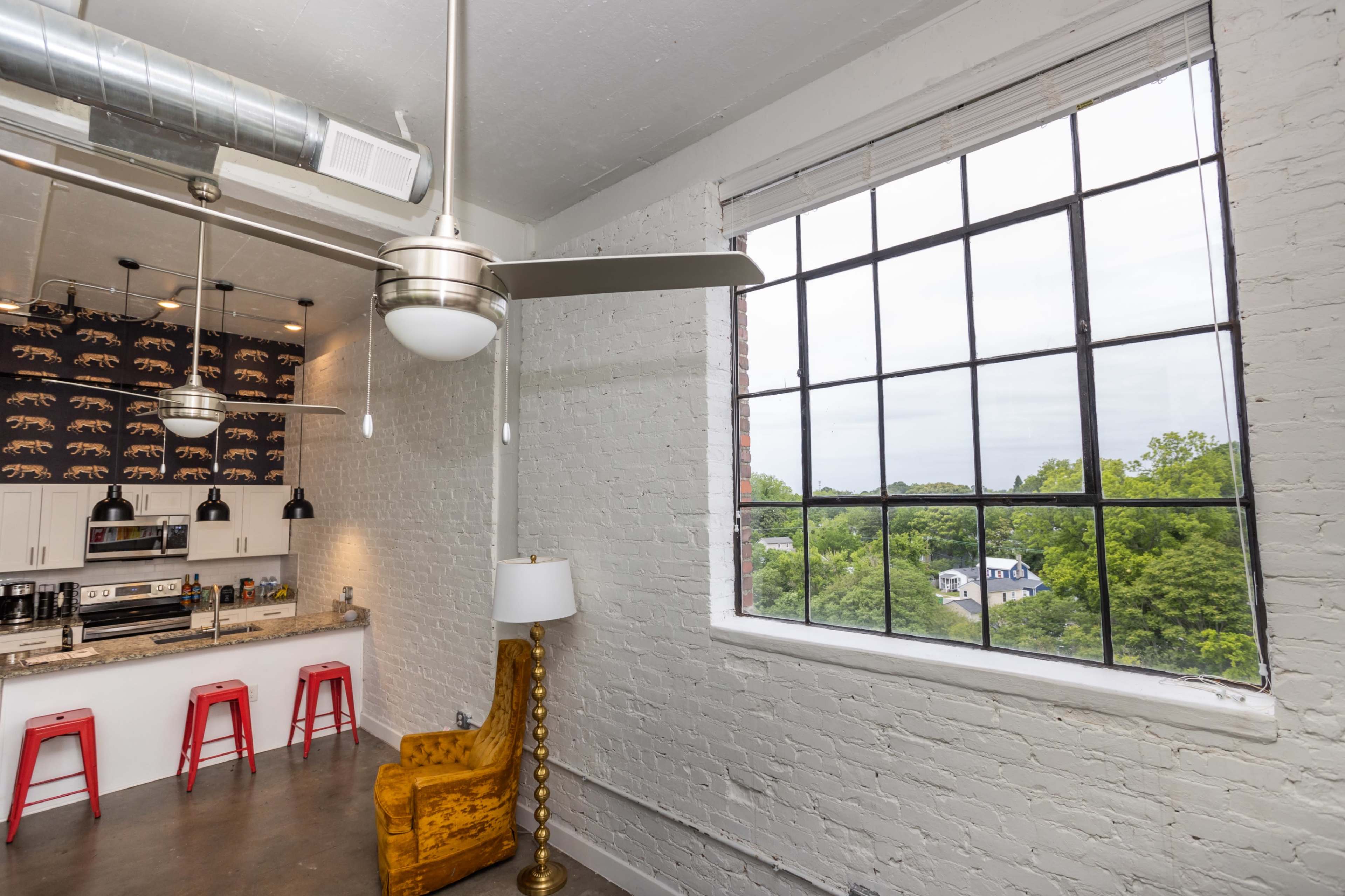 A modern kitchen area with red stools, a stylish lamp, and a large window offering a view of green trees outside.