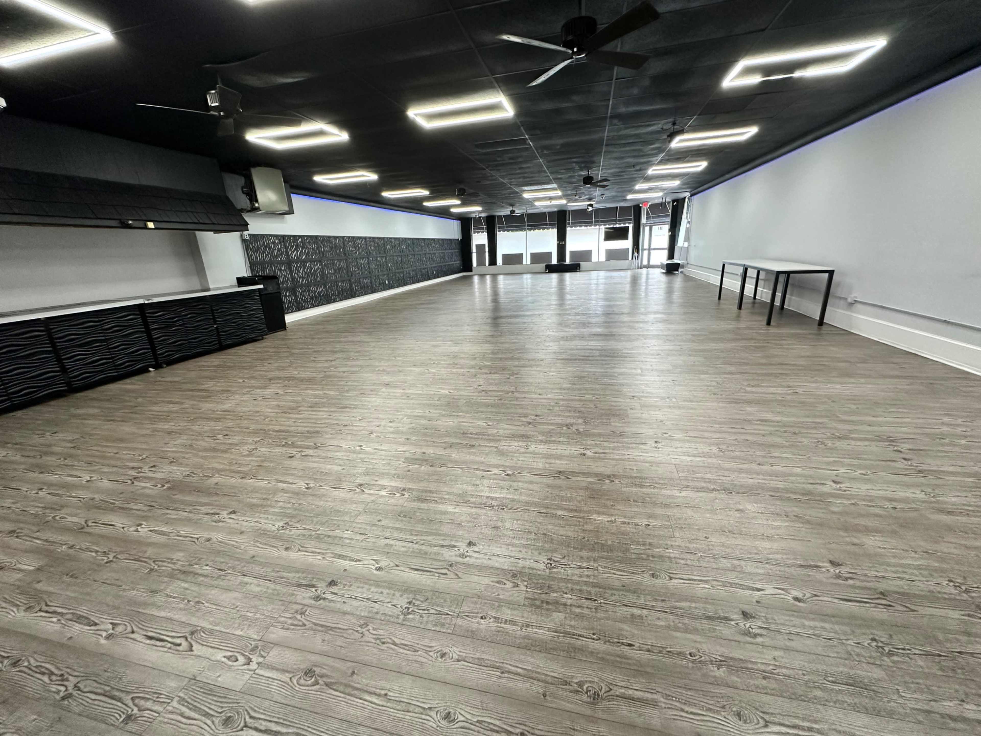 An empty interior space with wooden flooring, black ceiling fans, and a long, unobstructed layout featuring a front wall of large windows.