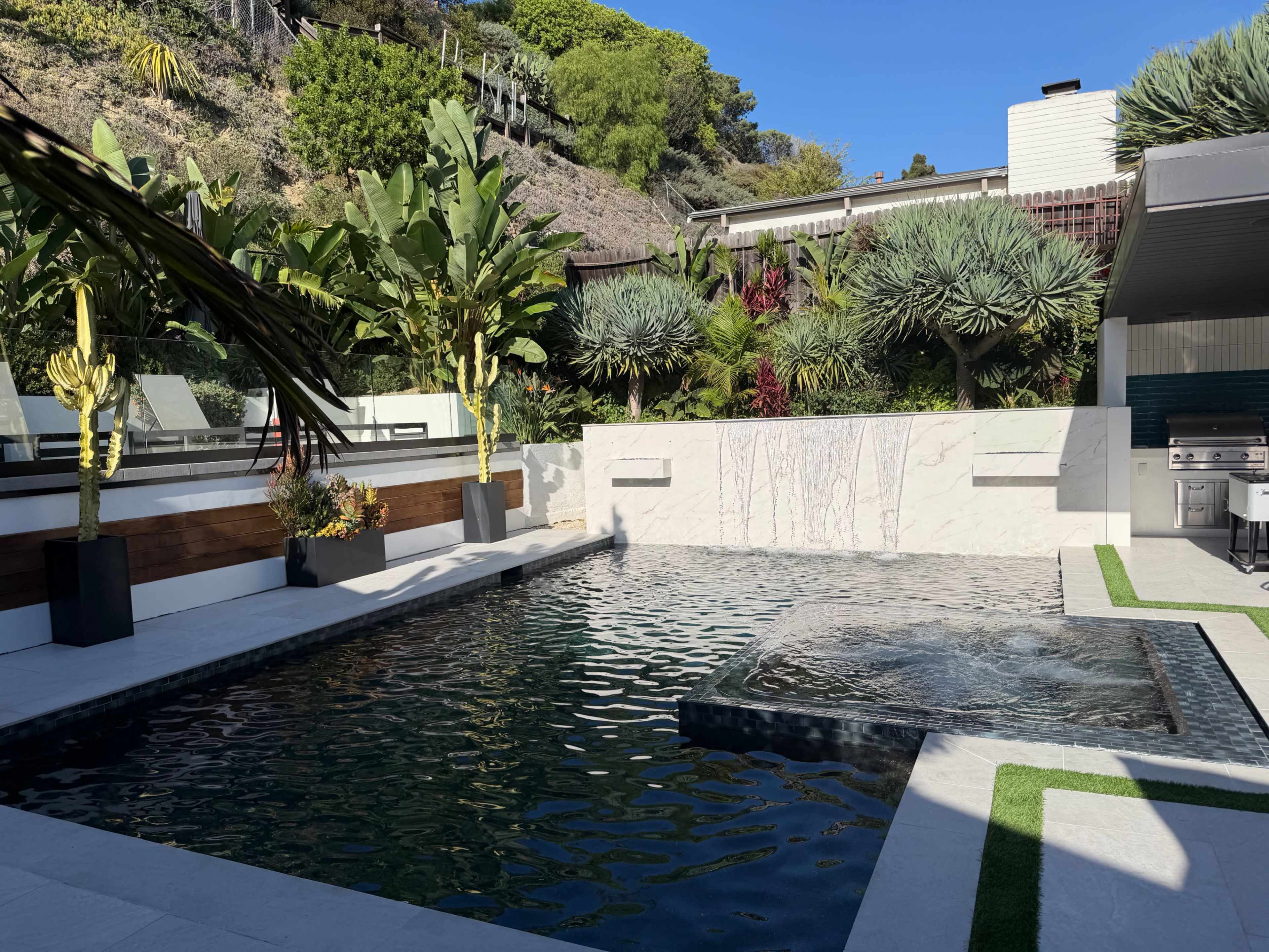 The image shows a modern outdoor space featuring a rectangular swimming pool with a jacuzzi, surrounded by tropical plants and stone pavement.