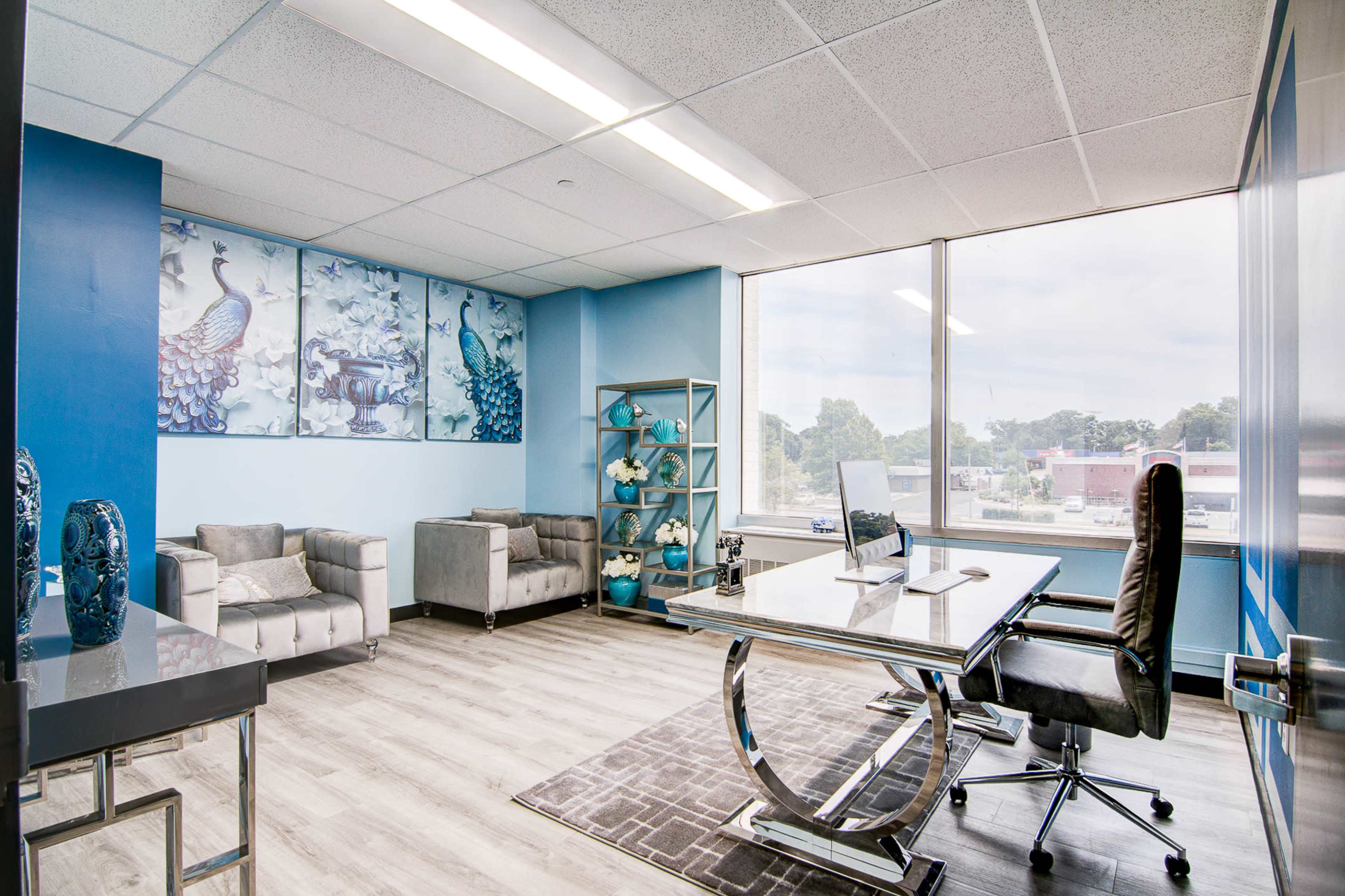 The image shows a modern office space featuring a glass desk, a black office chair, two gray sofas, and blue wall decor above a shelving unit.