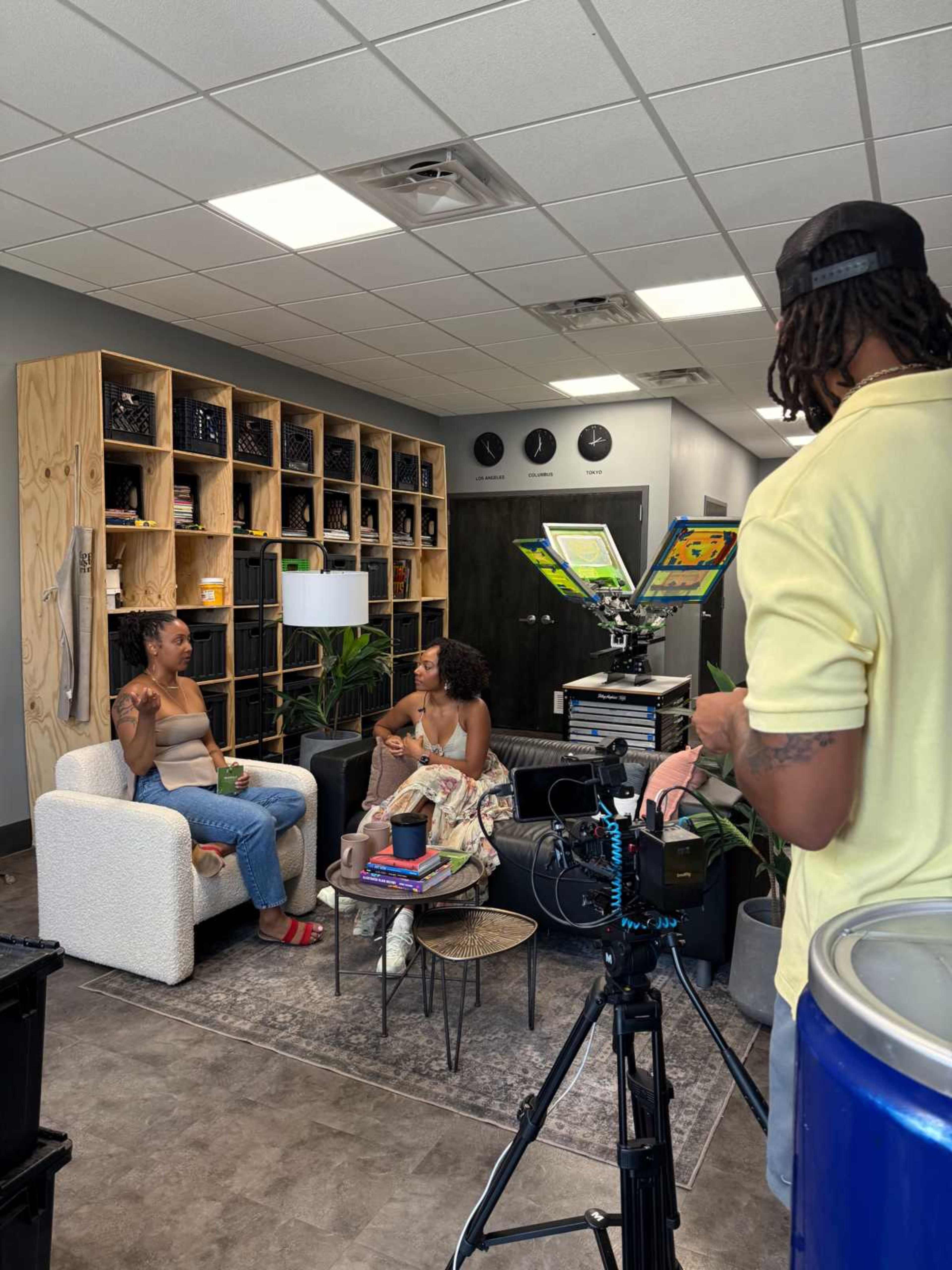 Two women are seated on a white couch engaged in conversation while a man sets up a camera in a well-furnished office space.