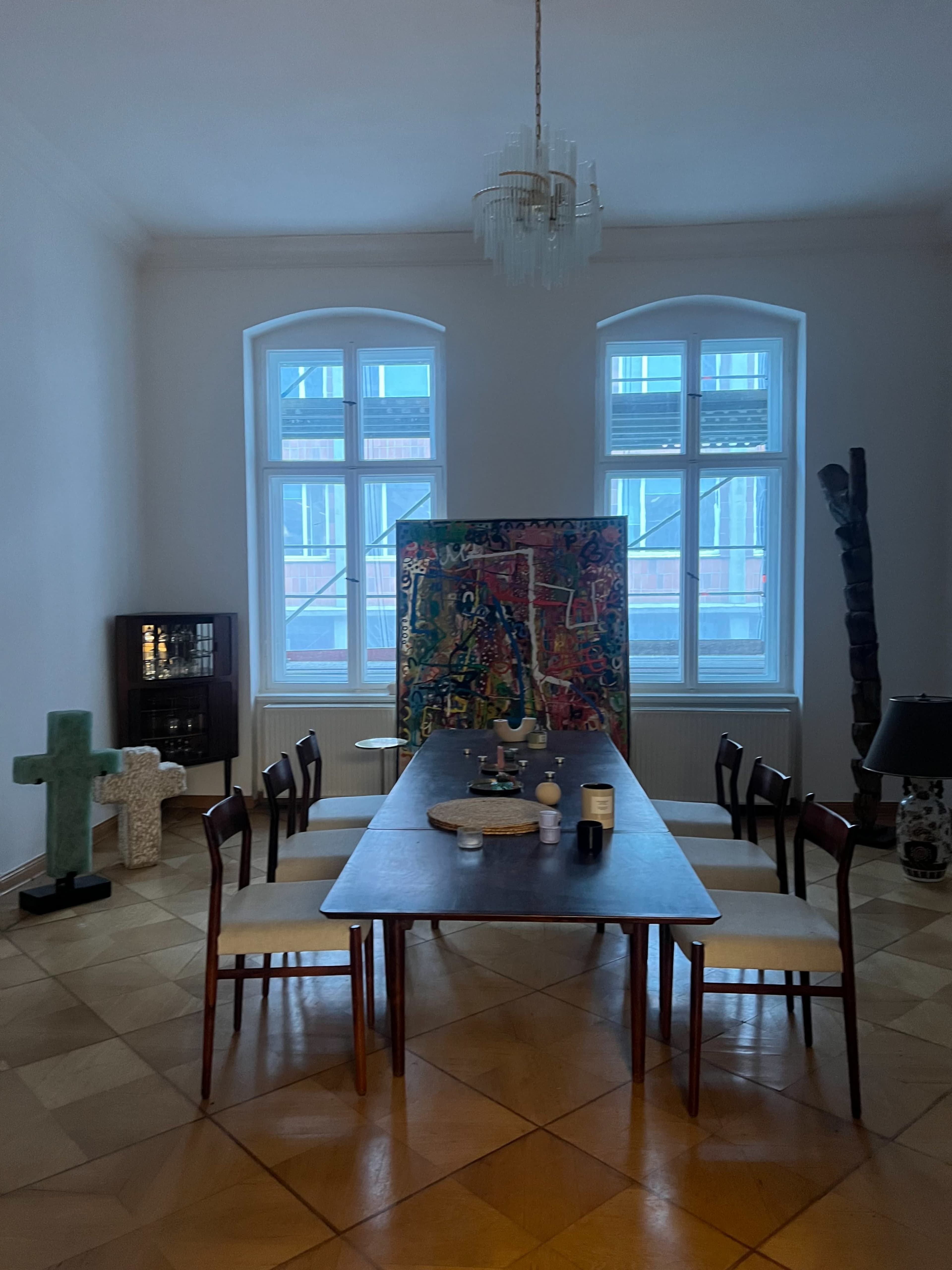 A long wooden dining table with several chairs is set in a room featuring large windows, a colorful abstract painting, and various decorative objects.
