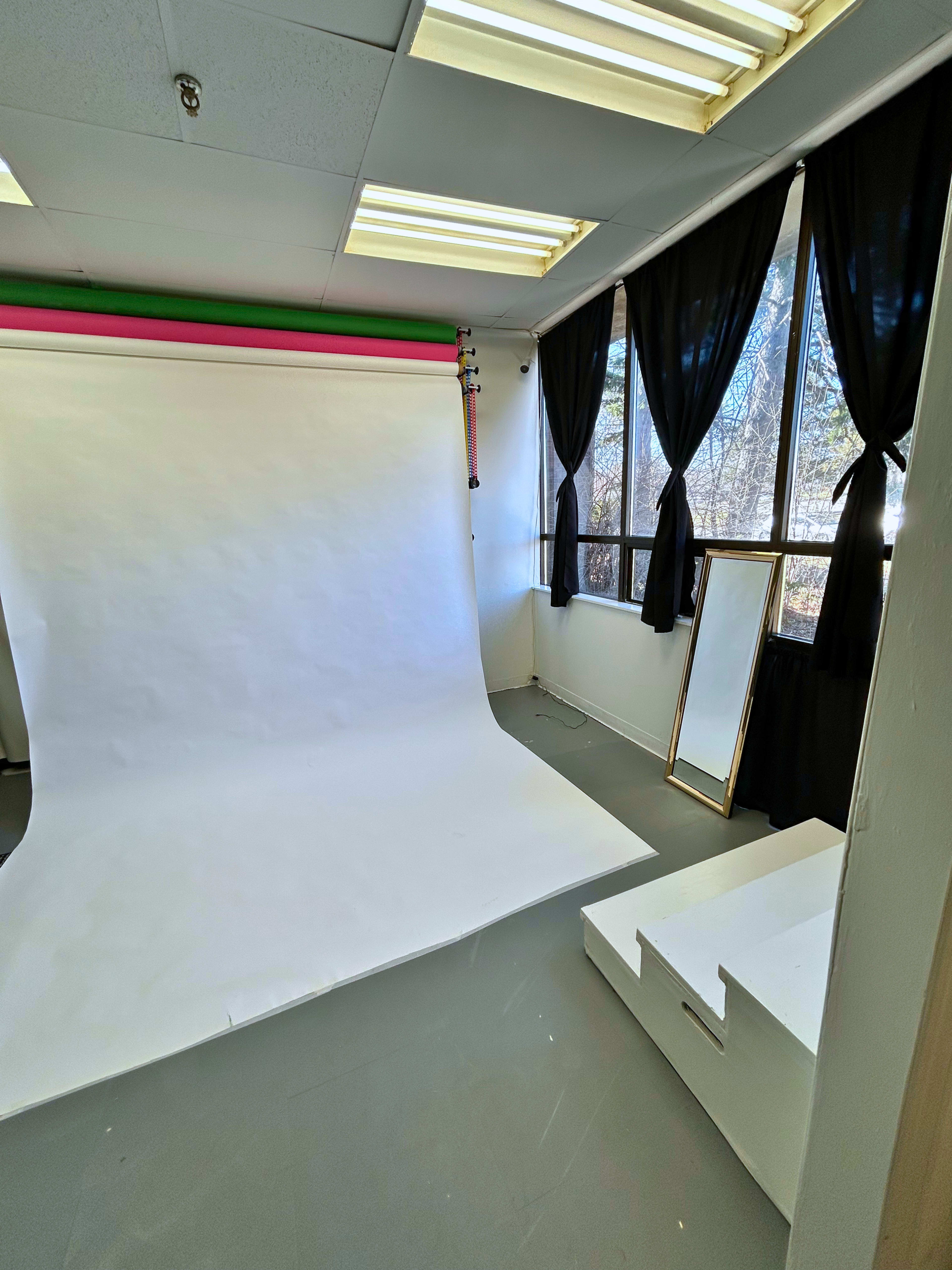 The image shows a brightly lit photo studio featuring a large white backdrop, a full-length mirror, and a small white storage unit, with large windows covered by black drapes.