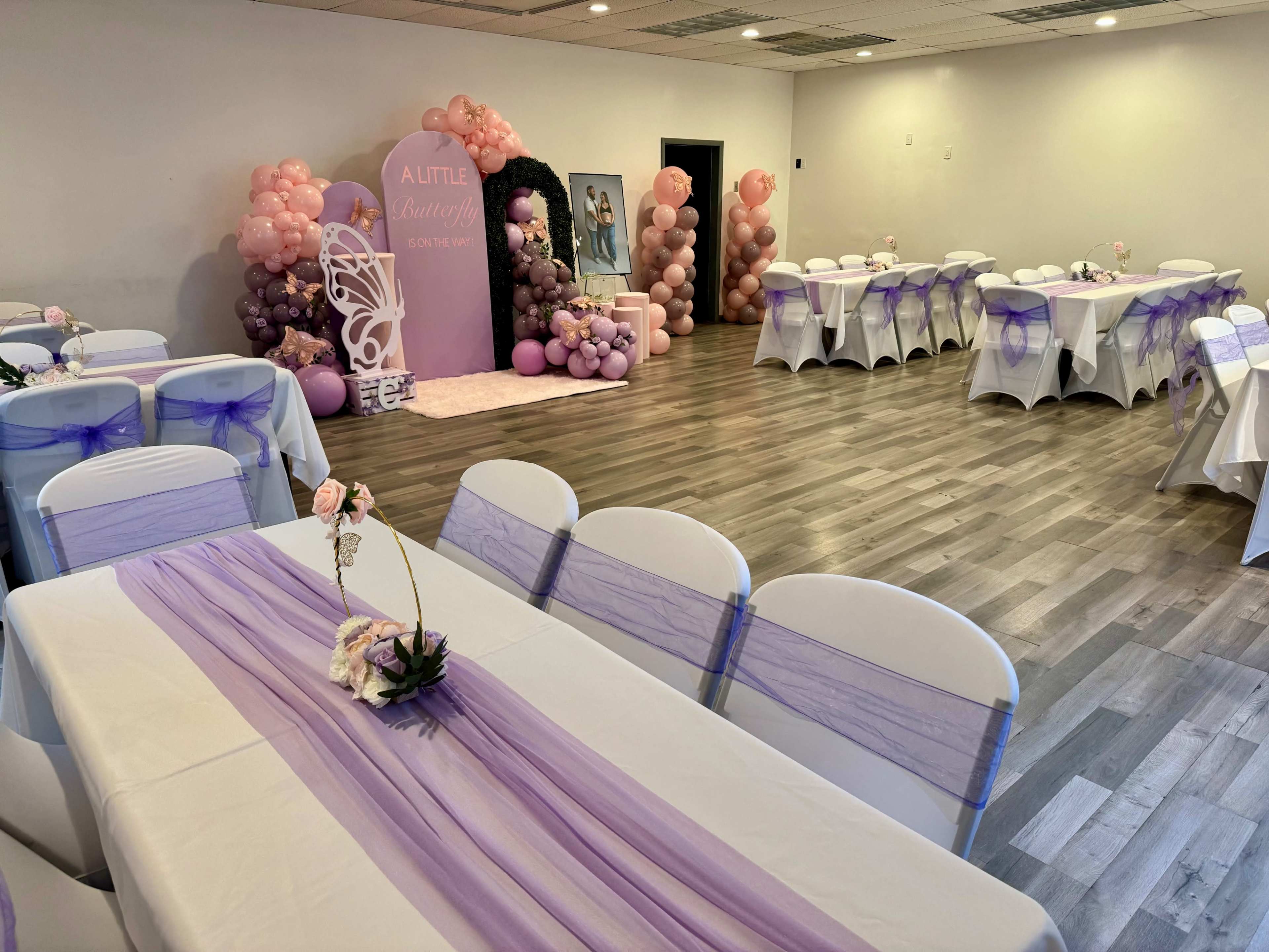 A decorated event space featuring white tables with purple table runners, surrounded by balloon arrangements and a backdrop for a celebration.