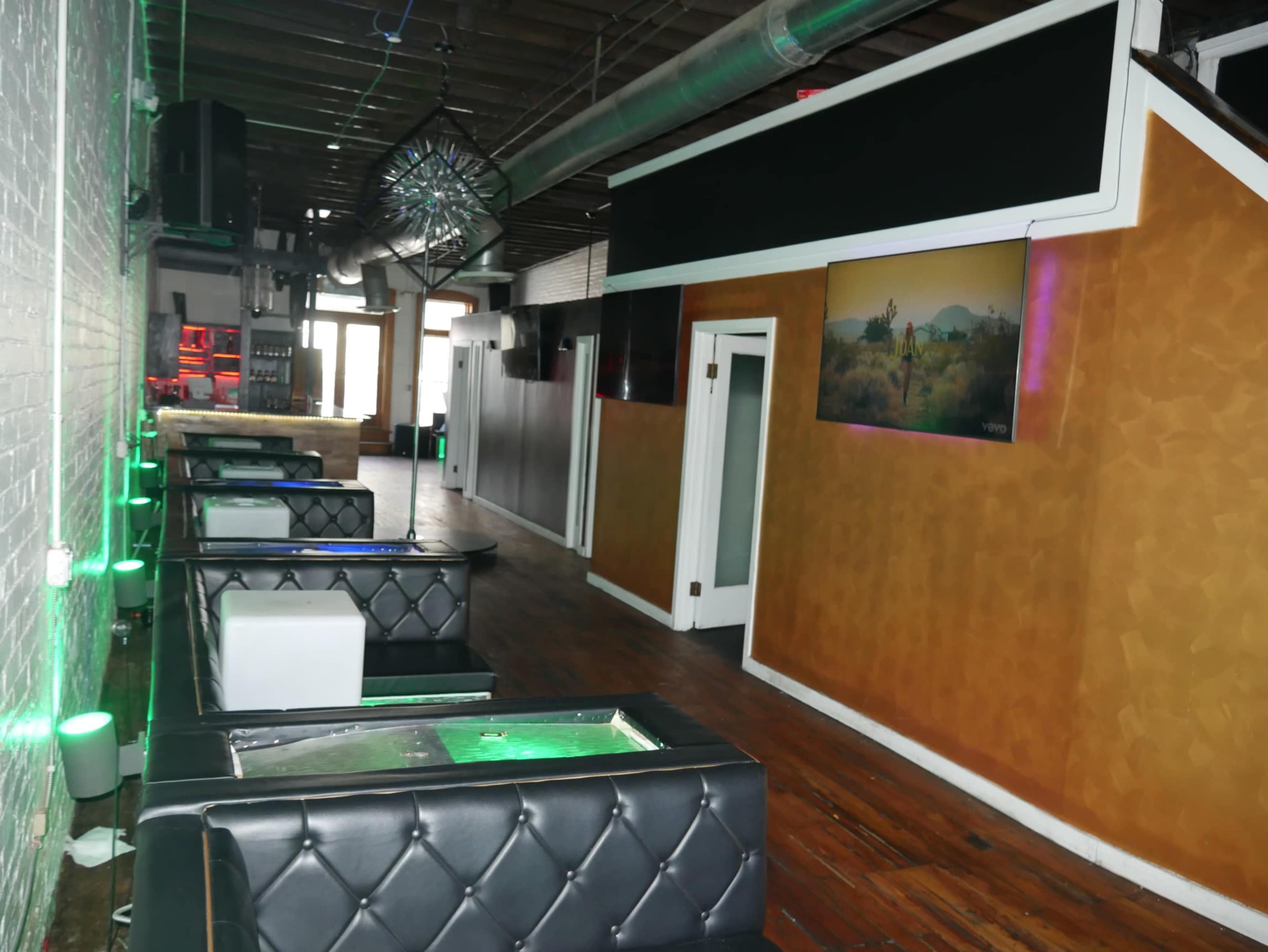 An empty lounge or bar area with leather seating and green-lit tables along a wall, leading to a doorway and a large window at the far end.