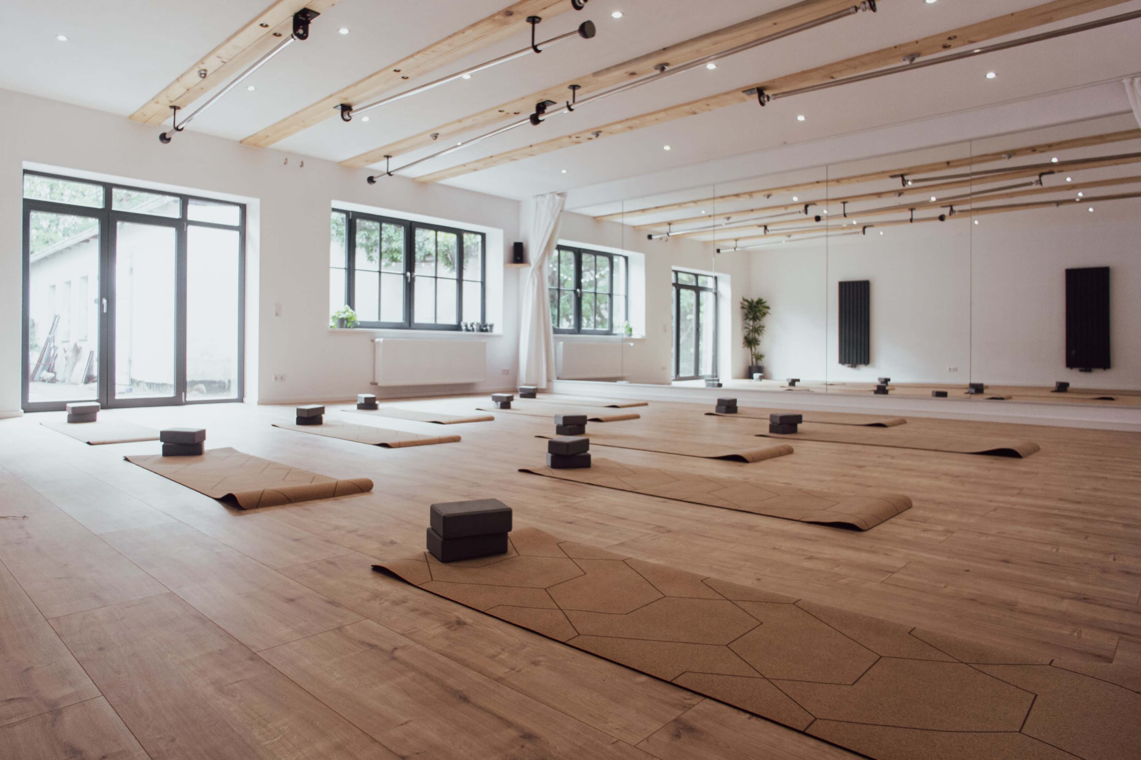 The image shows a spacious yoga studio with several mats and blocks organized on the wooden floor, surrounded by large windows and bright lighting.