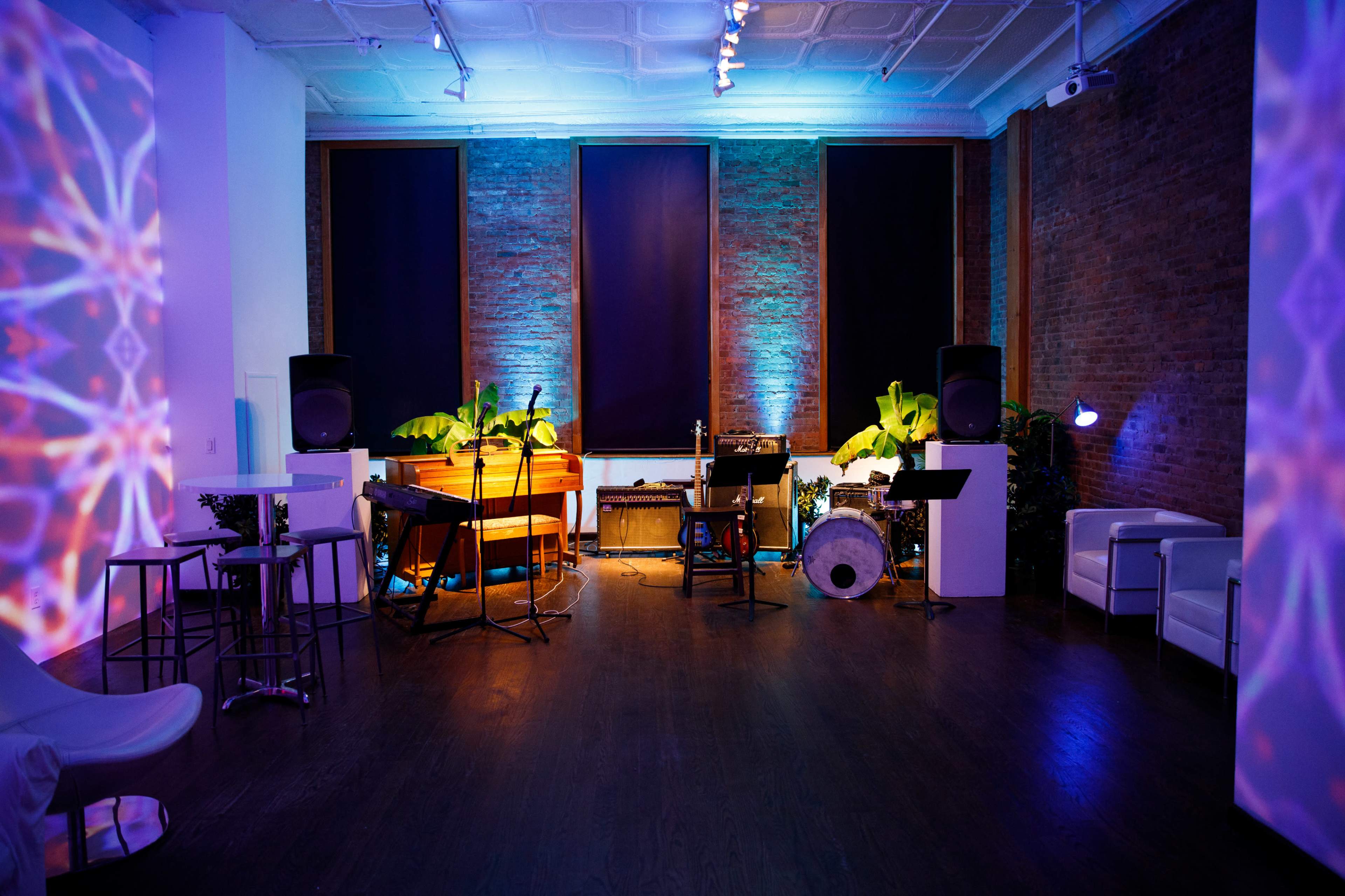 A dimly lit venue featuring a stage with musical instruments, chairs, and decorative plants against a brick wall.