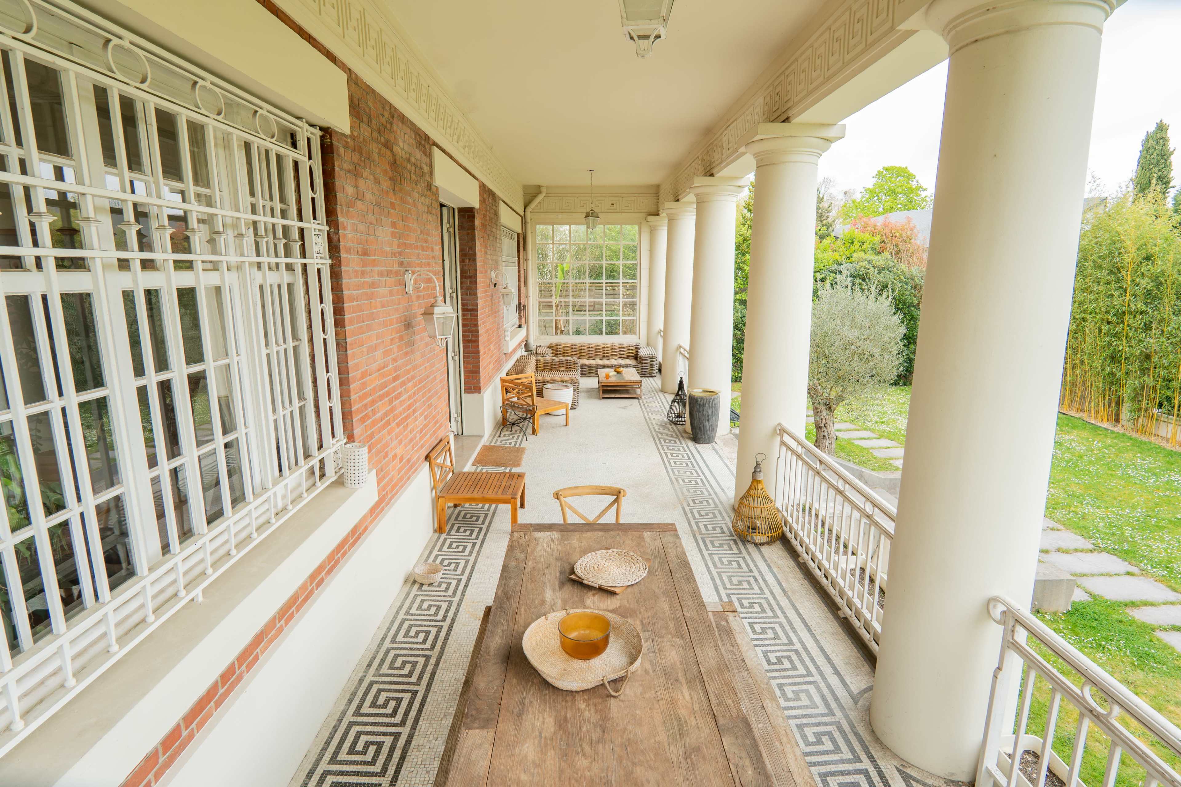 The image shows a covered patio with a wooden dining table, surrounded by columns, chairs, and greenery in the background.