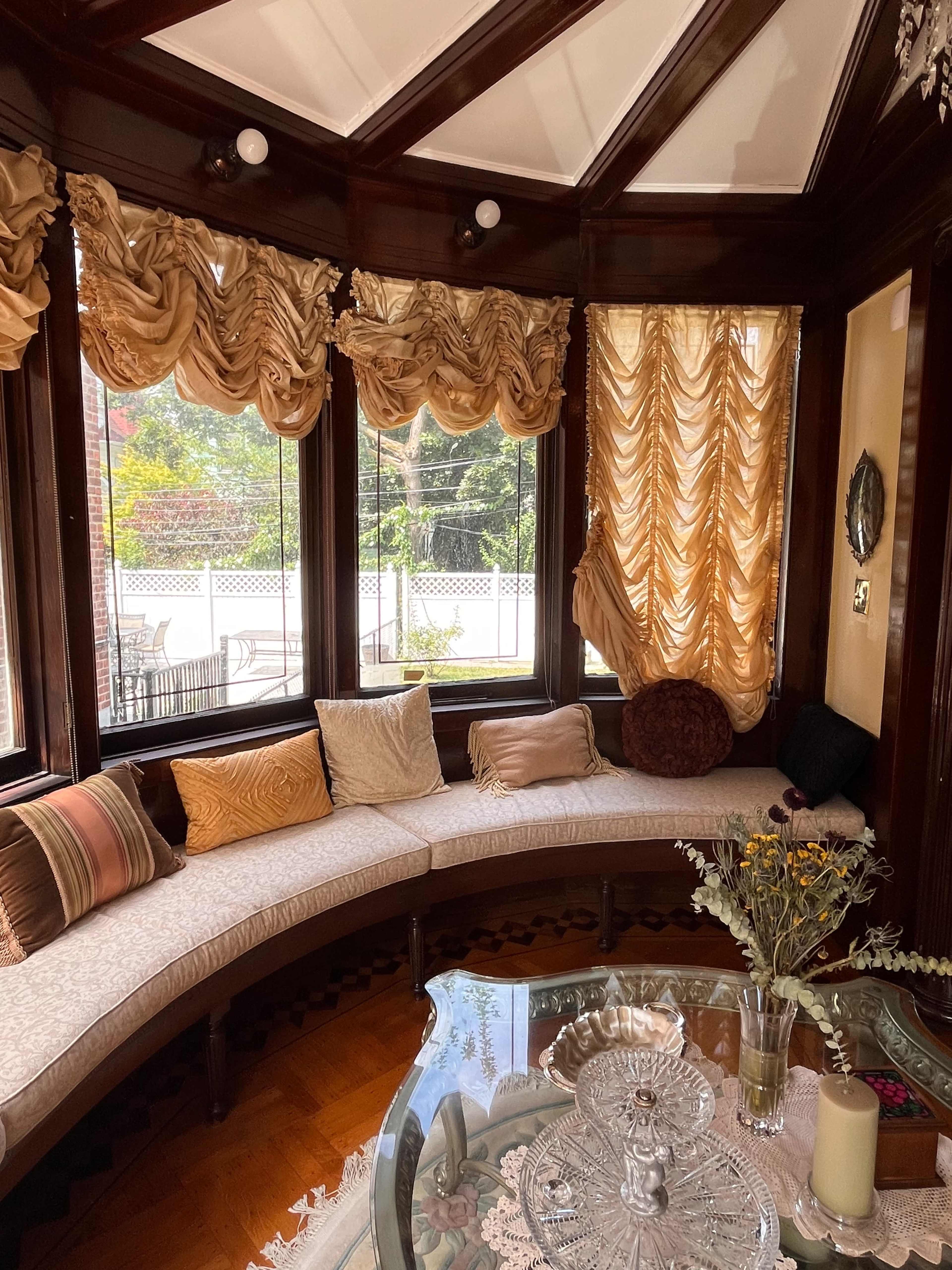 A cozy sunroom features a curved seating area with decorative pillows, large windows draped with golden curtains, and a glass coffee table topped with flowers and a candle.