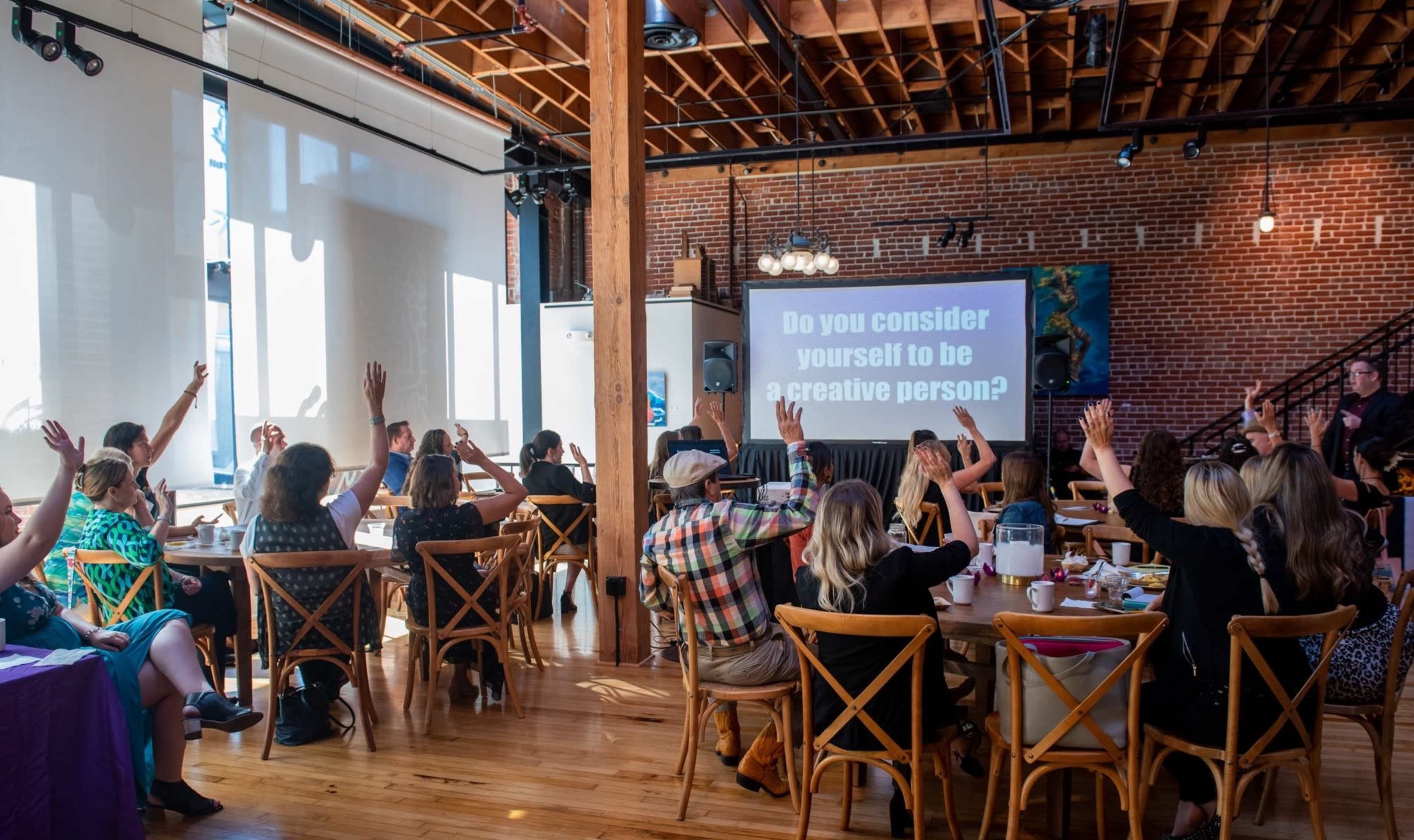 A group of people sits at tables in a spacious room with brick walls, raising their hands in response to a question displayed on a screen.