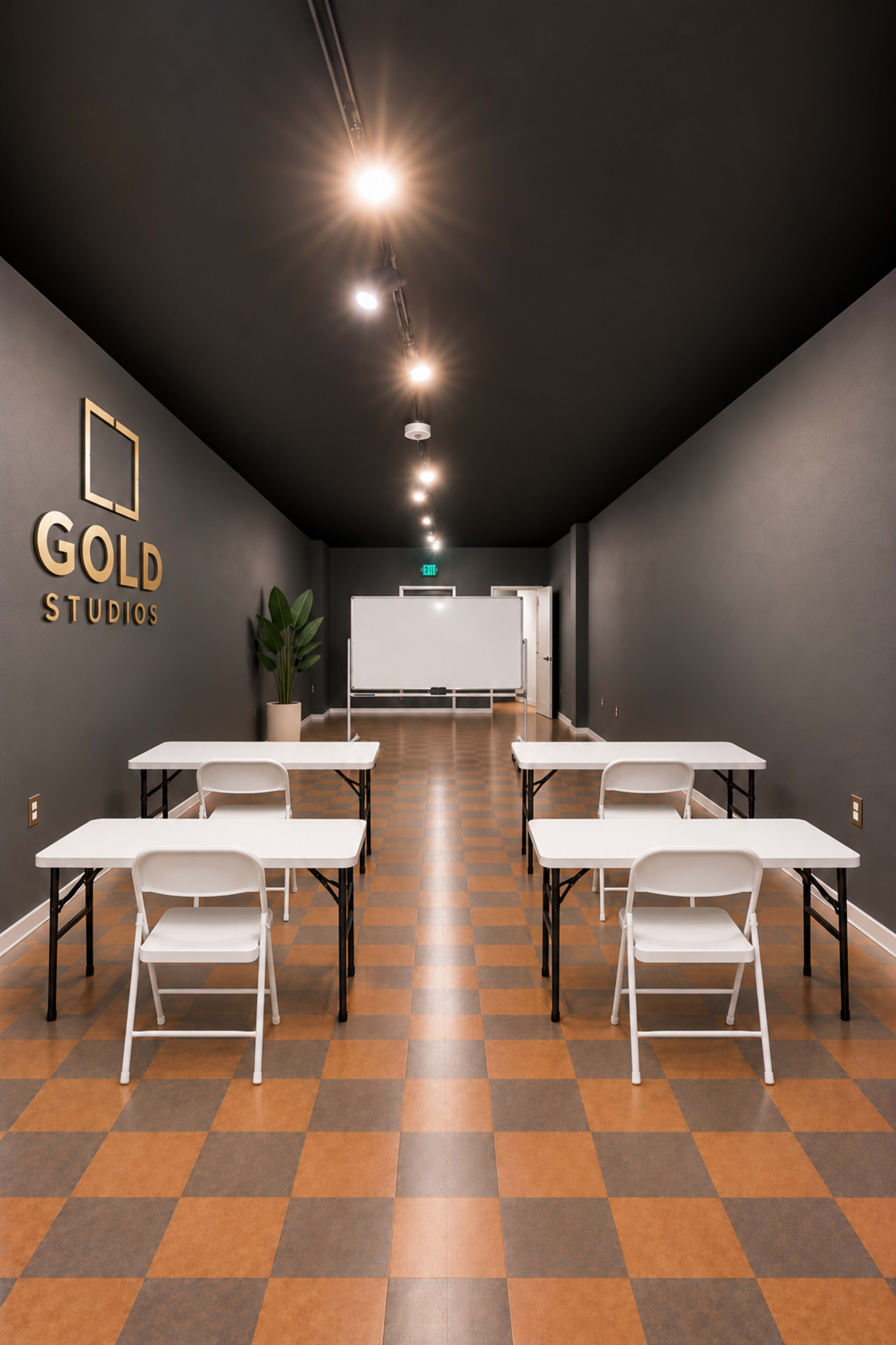 The image shows a long, narrow room with several white folding tables and chairs arranged in rows, a whiteboard at the far end, and a sign that reads "GOLD STUDIOS" on the wall.