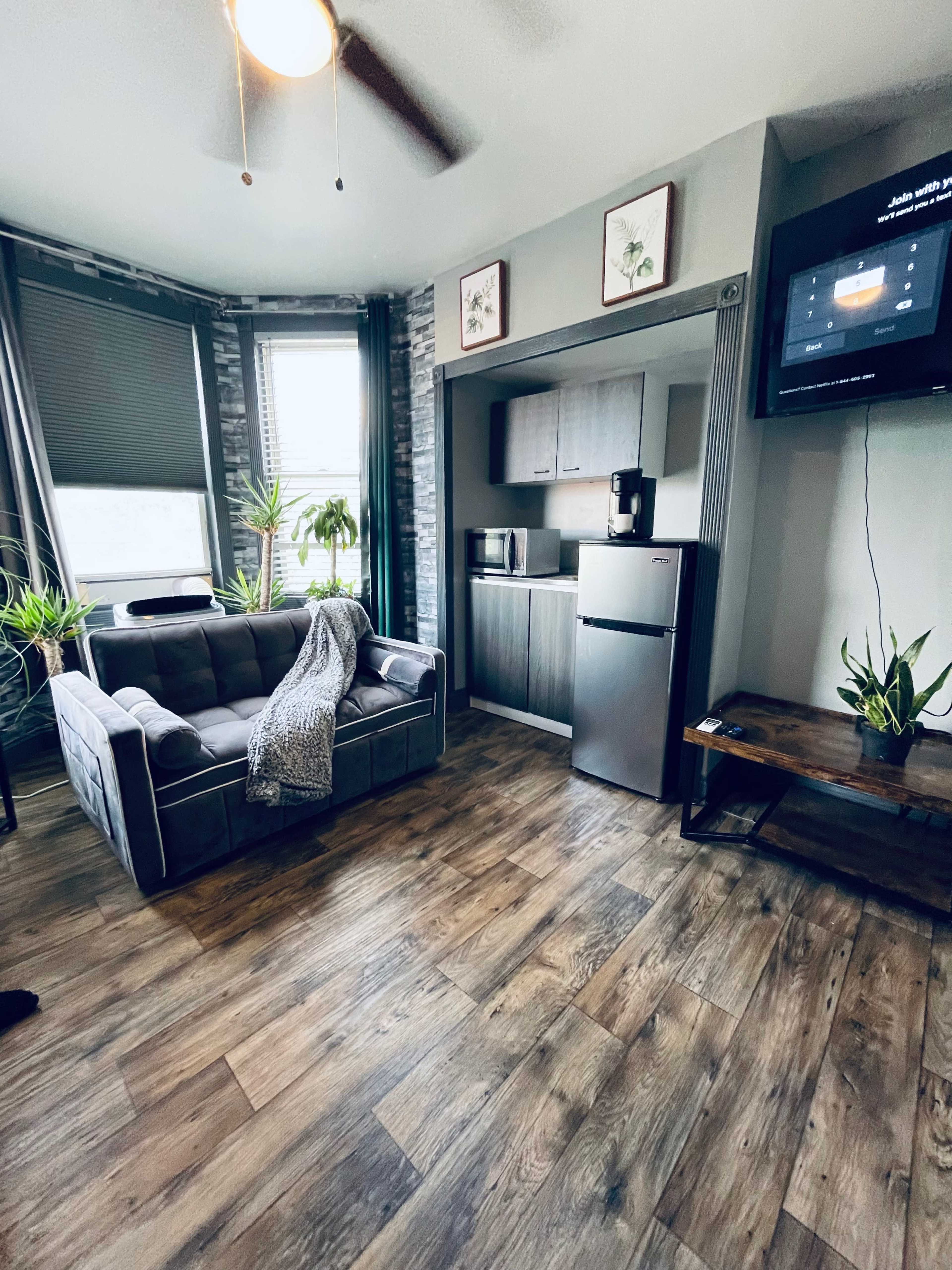 A modern living space featuring a gray sofa, a kitchenette with a refrigerator, and potted plants near large windows.