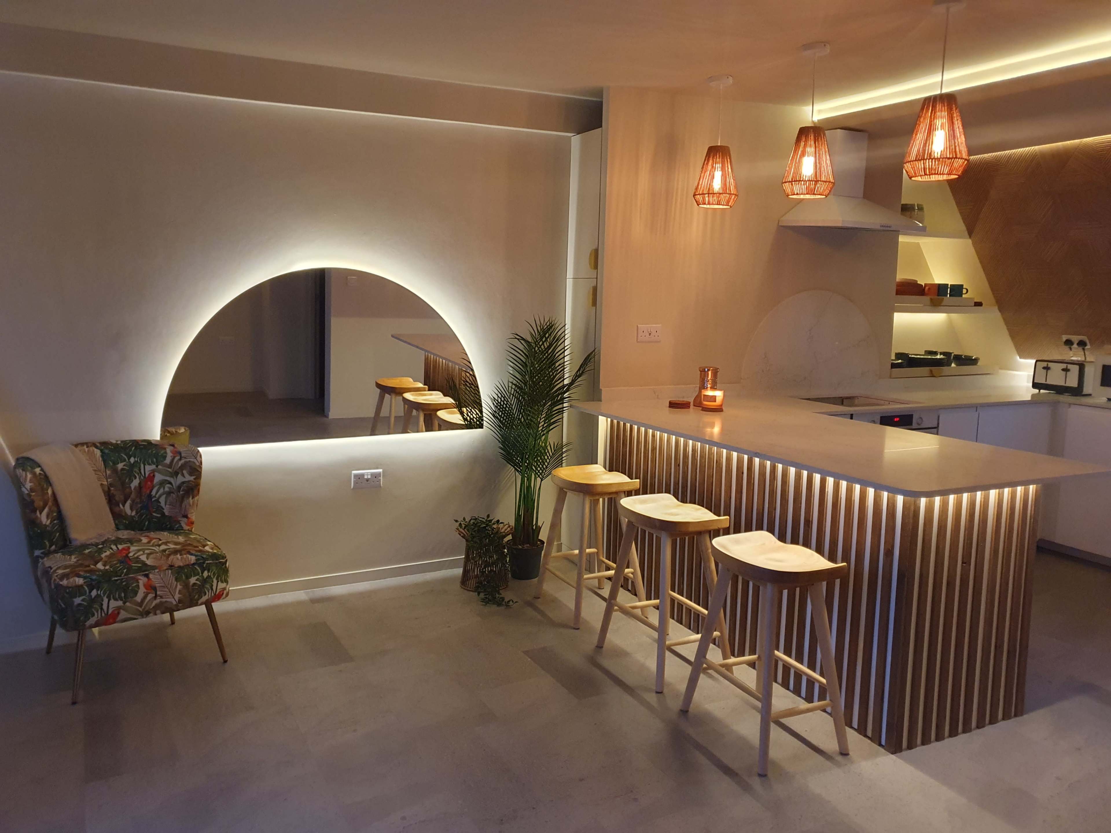 A modern kitchen with a curved mirror, wooden bar with stools, and pendant lights cast in warm tones.