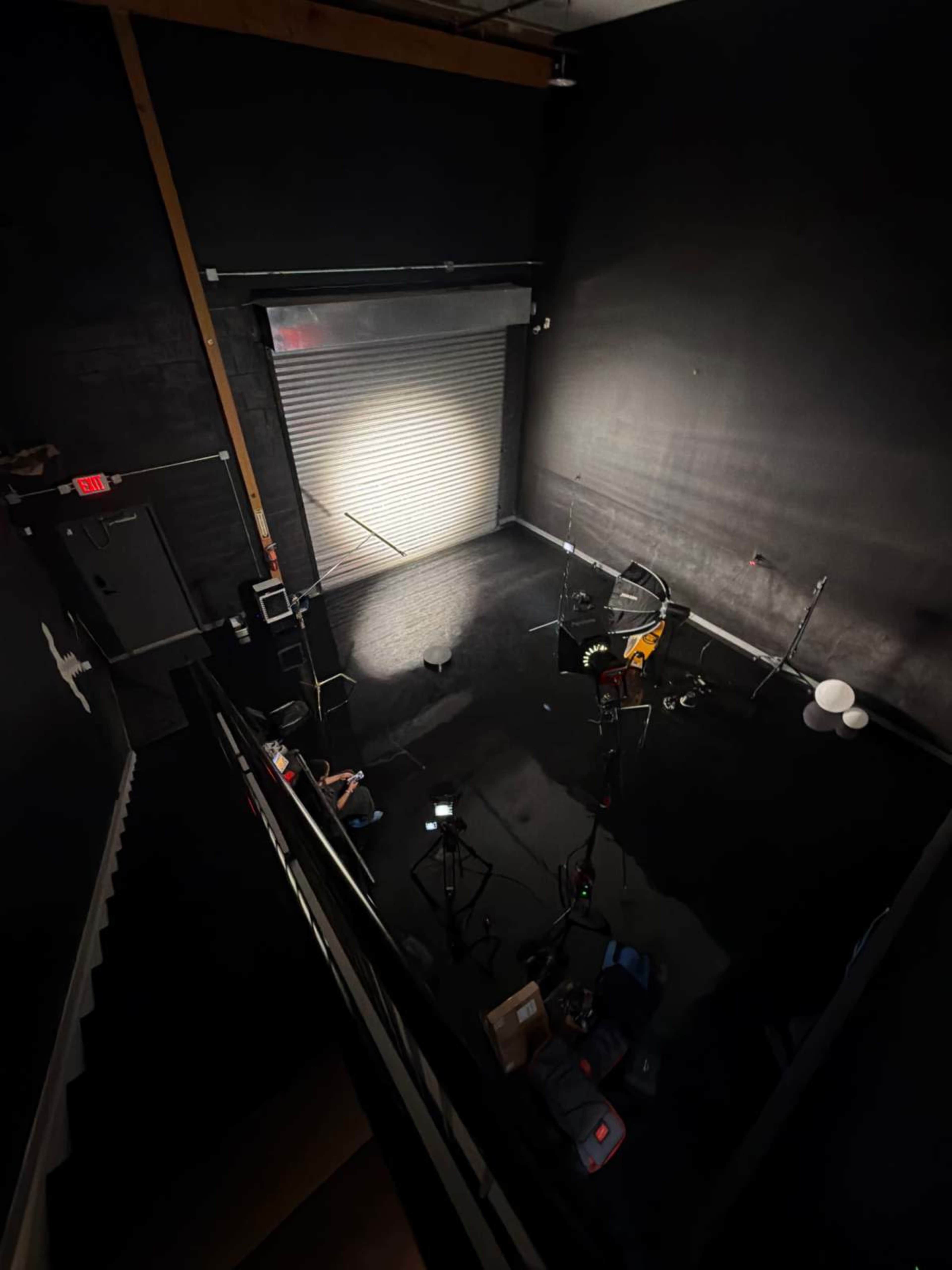 The image shows an empty black room with a guitar and microphone set up in front of a closed garage door, illuminated by a single spotlight.