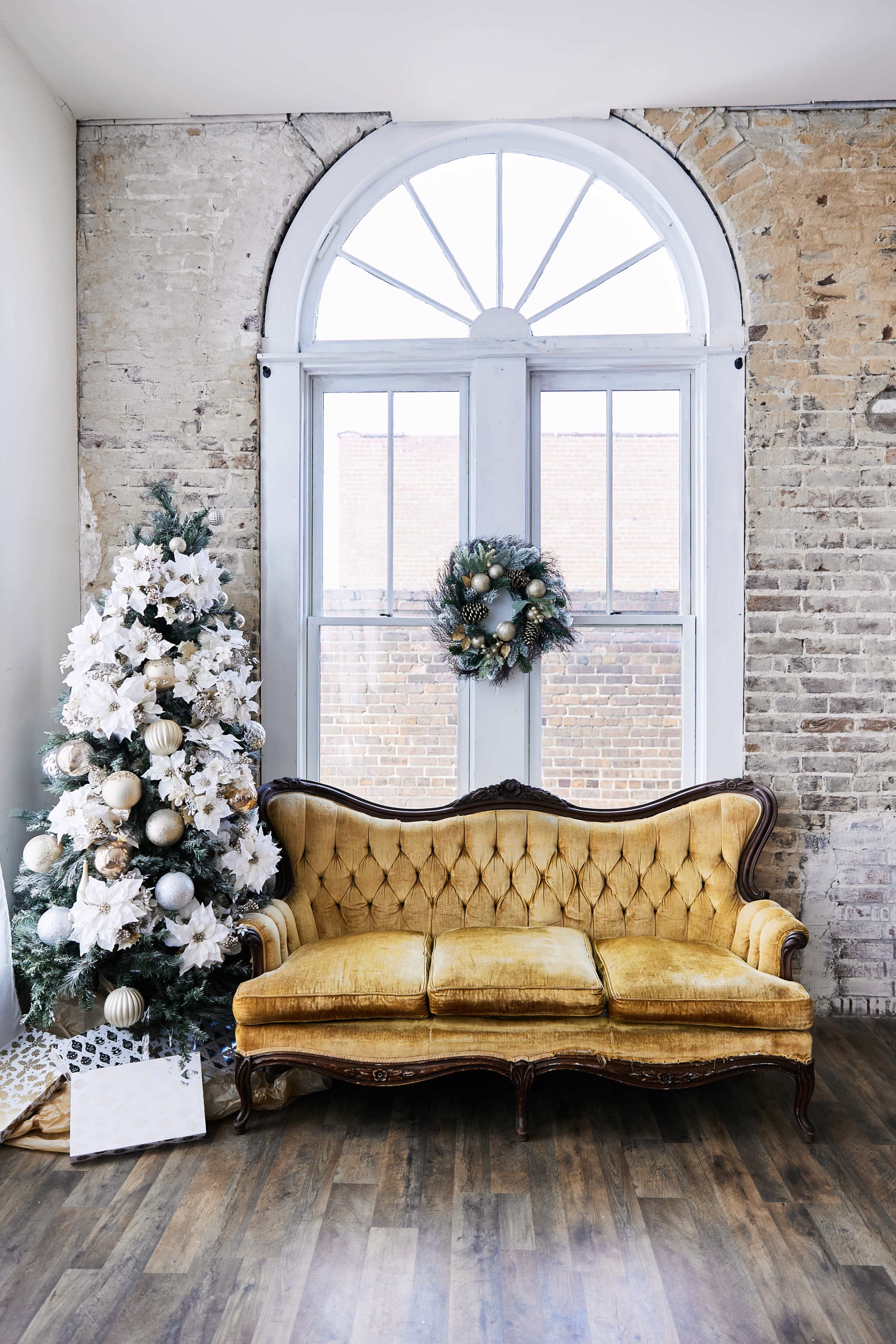 A vintage yellow tufted sofa is positioned in front of a large window, with a decorated Christmas tree and a wreath nearby.