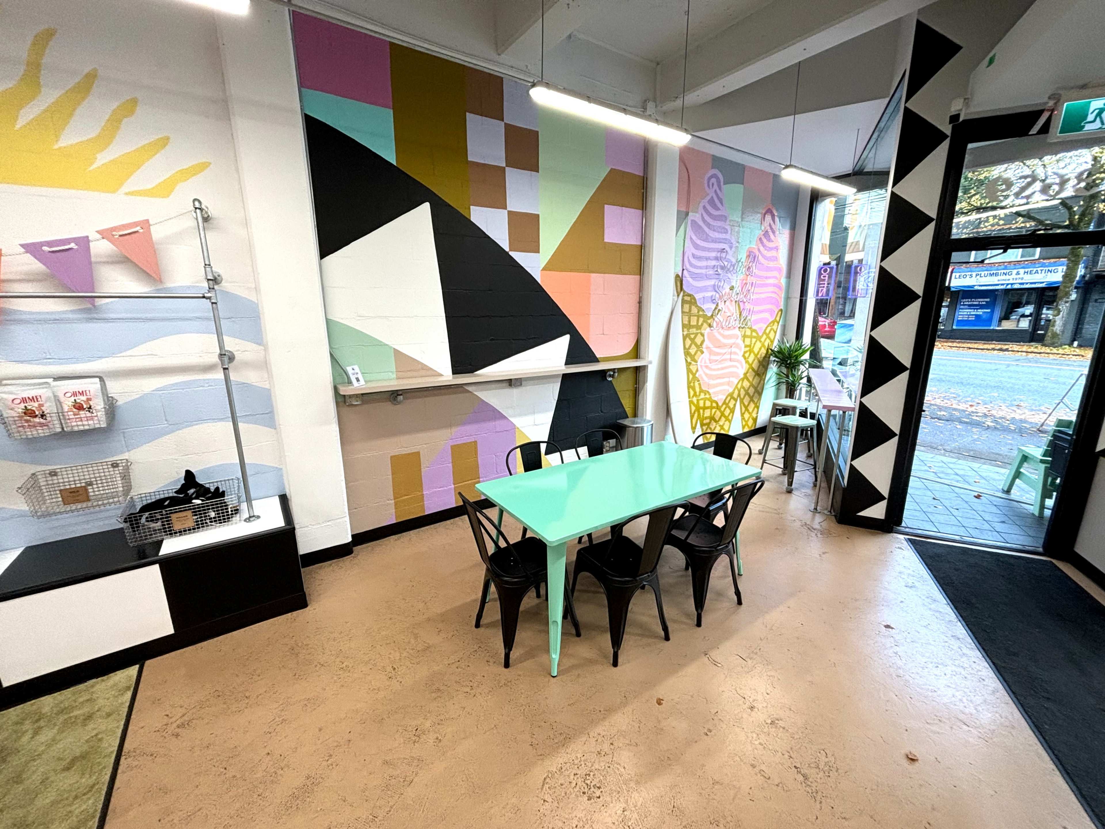 The image shows a modern cafe interior with colorful abstract wall murals, a mint green table, black chairs, and large windows facing the street.