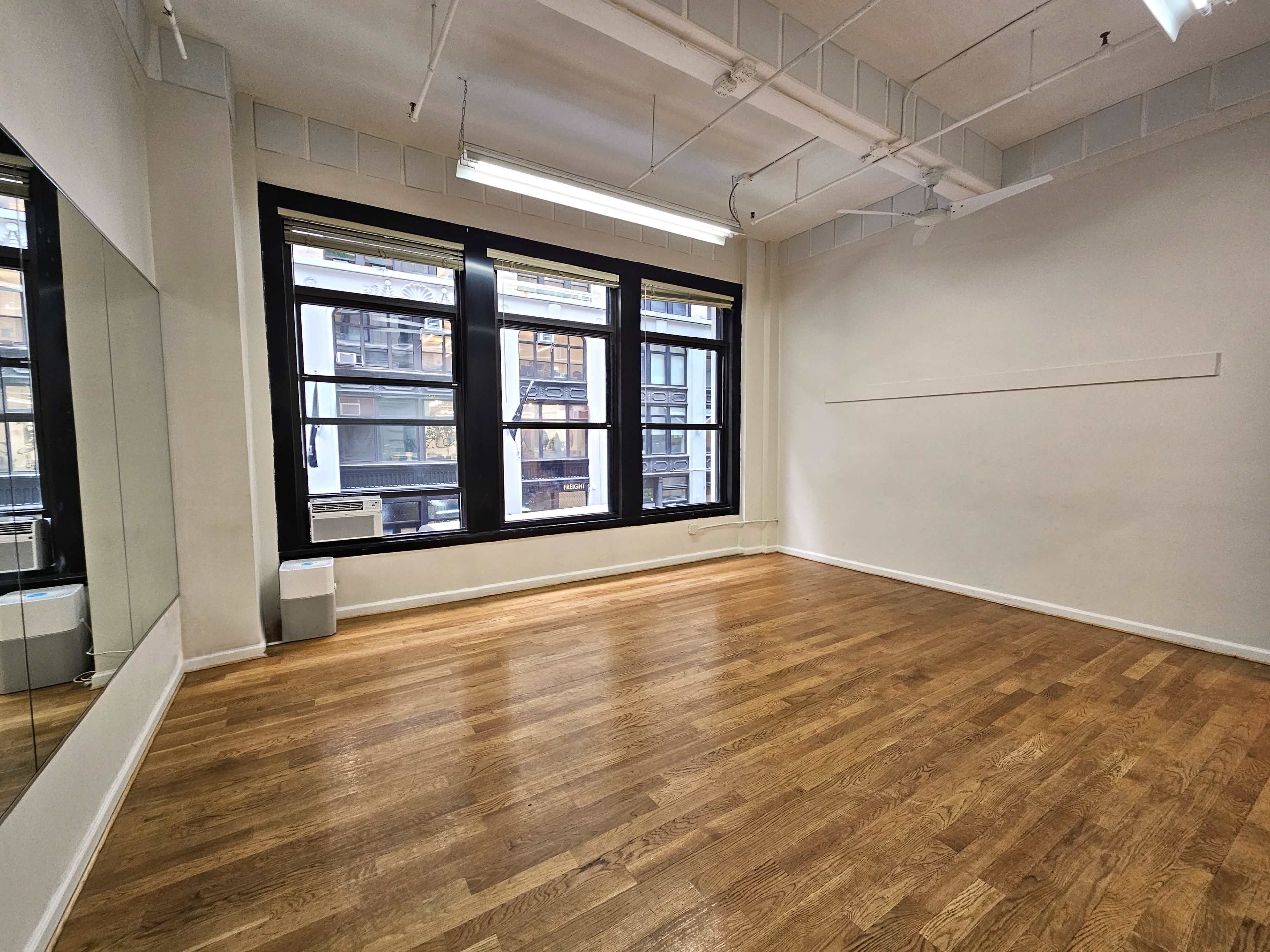 An empty room with hardwood flooring, large windows on one side, and a mirror on the wall opposite the windows.
