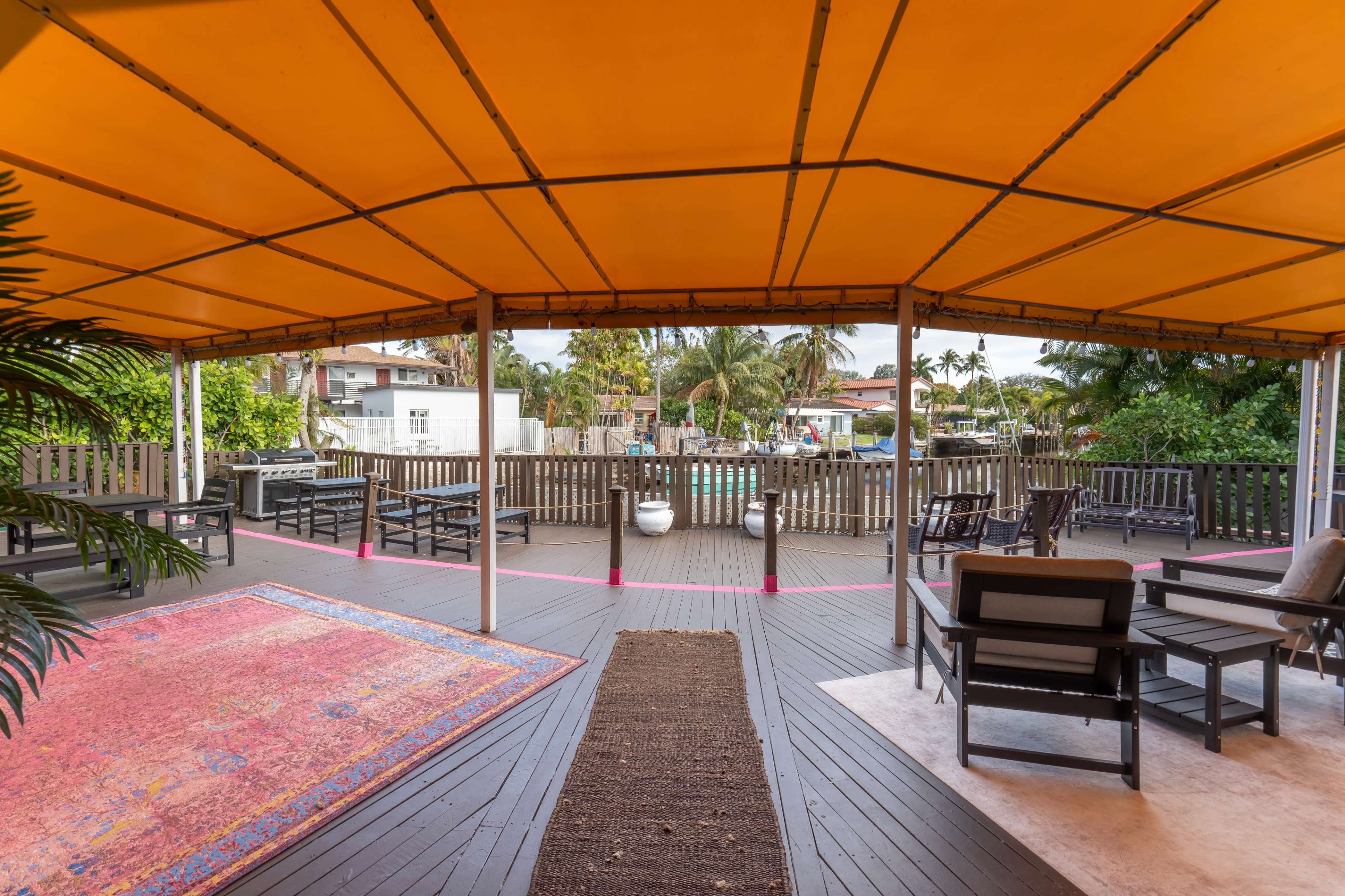 The image shows a spacious outdoor patio area with an orange canopy, wooden furniture, and a view of a pool surrounded by palm trees.