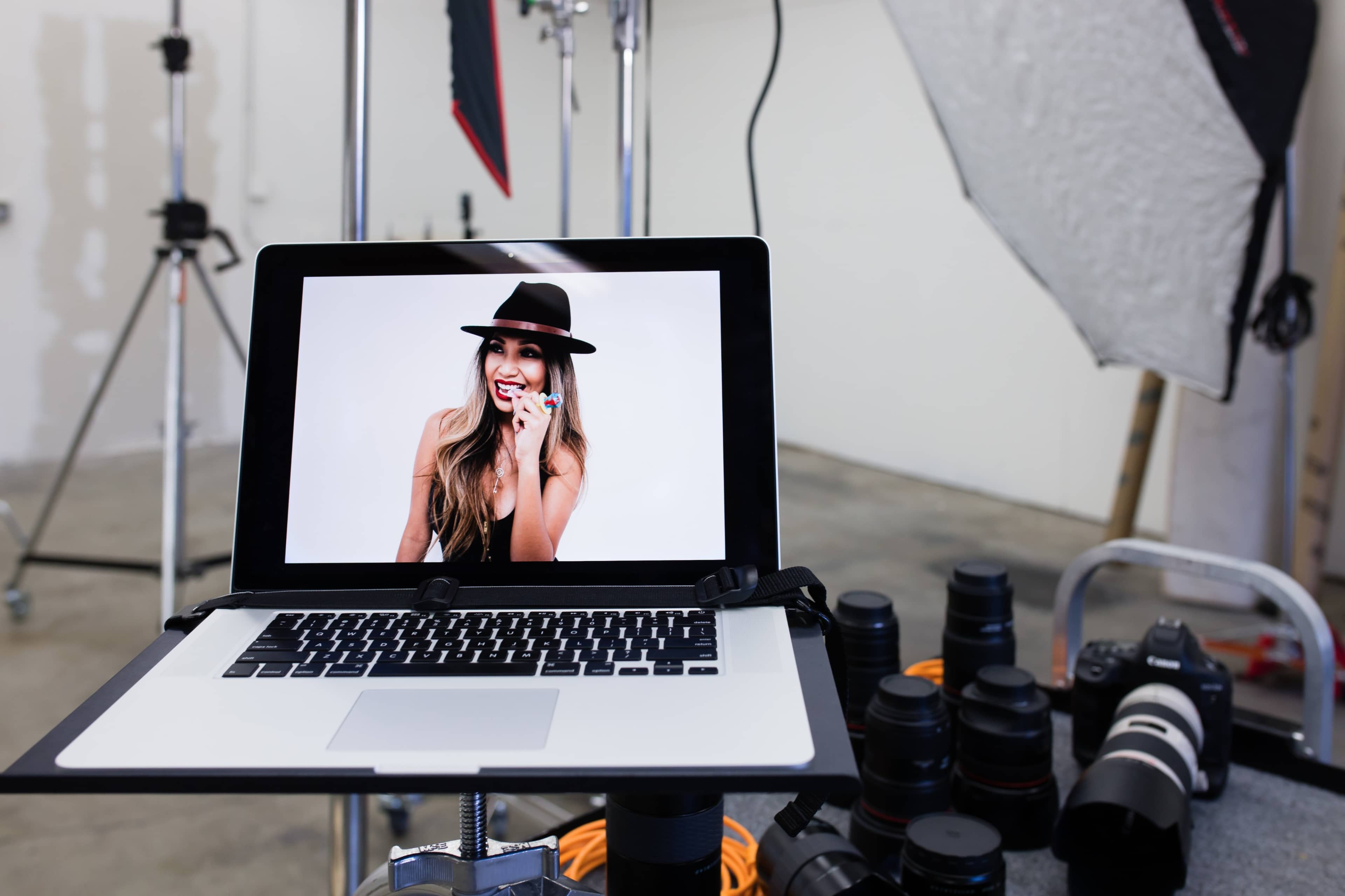A laptop displaying a model smiling is set up on a table next to various camera lenses and lighting equipment in a photography studio.