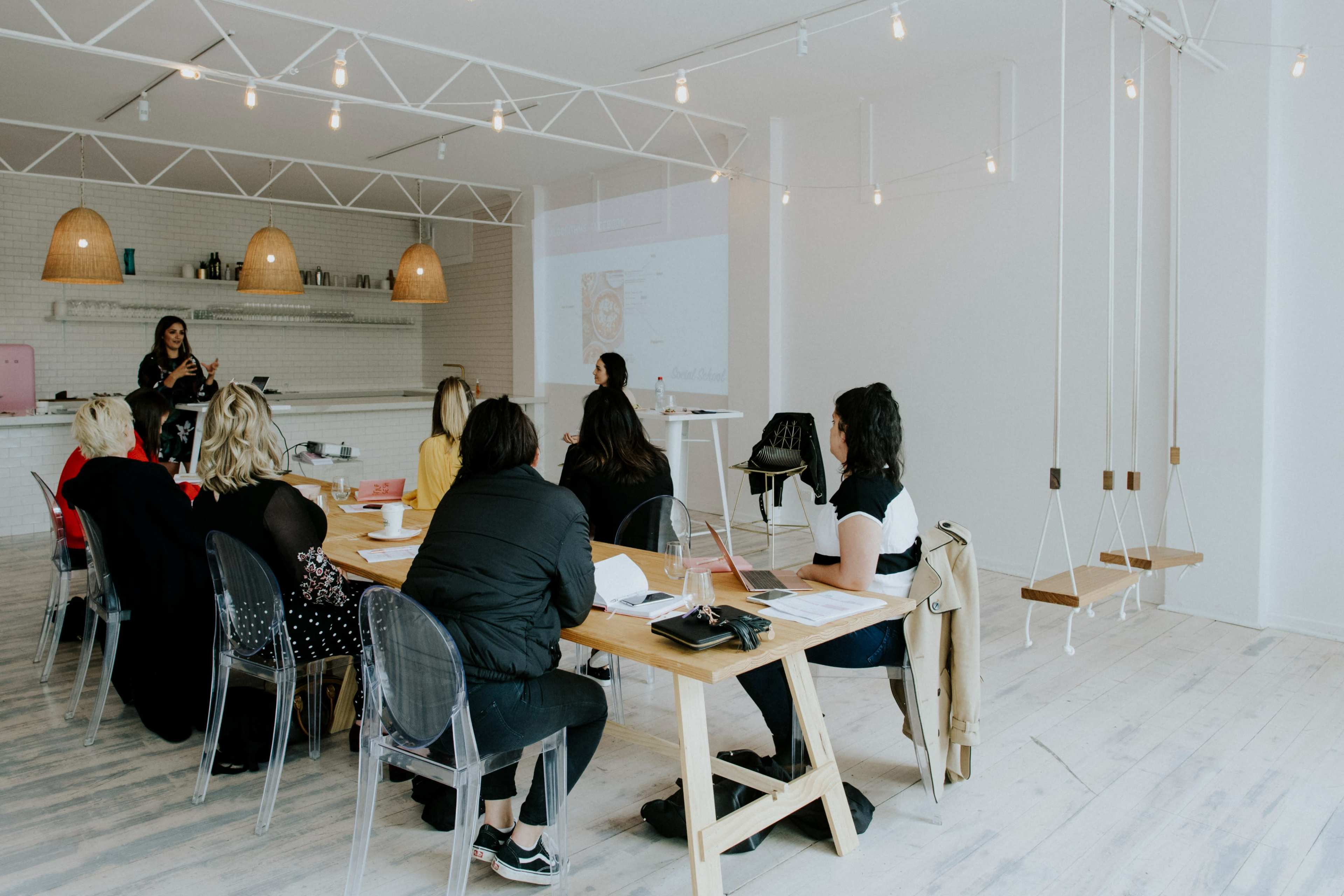A group of people is sitting around a long wooden table in a bright, minimalistic room, watching a presentation on a screen.