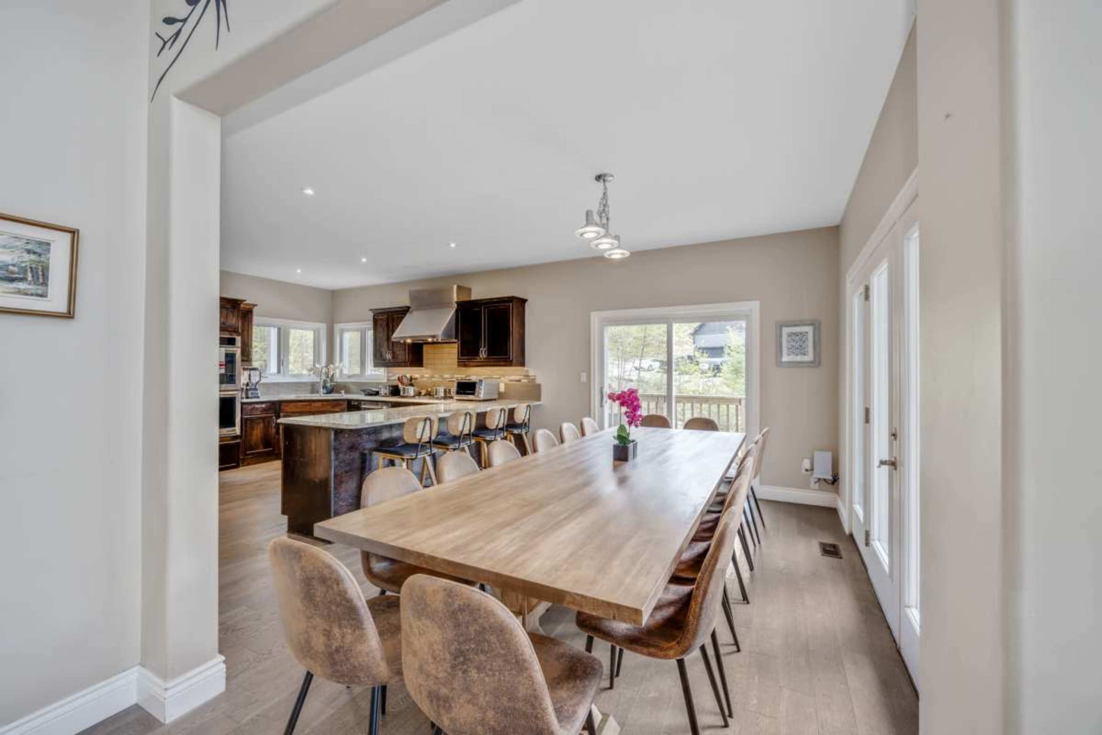 A large wooden dining table with chairs is situated in a bright dining area that opens into a modern kitchen.