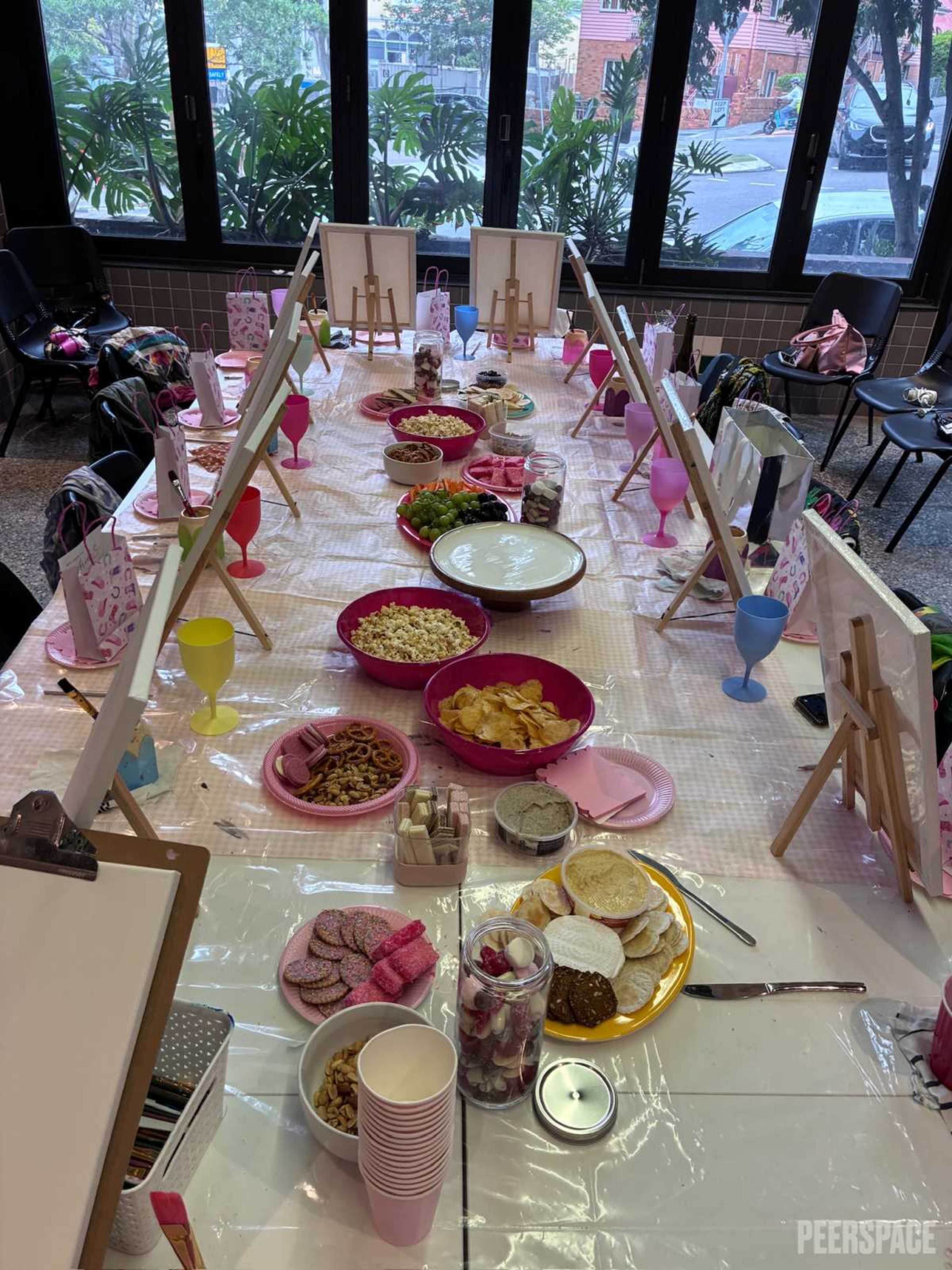 A long table is set for a gathering, featuring various dishes, snacks, and beverages organized in colorful bowls and cups, with empty canvases and easels placed at each side.