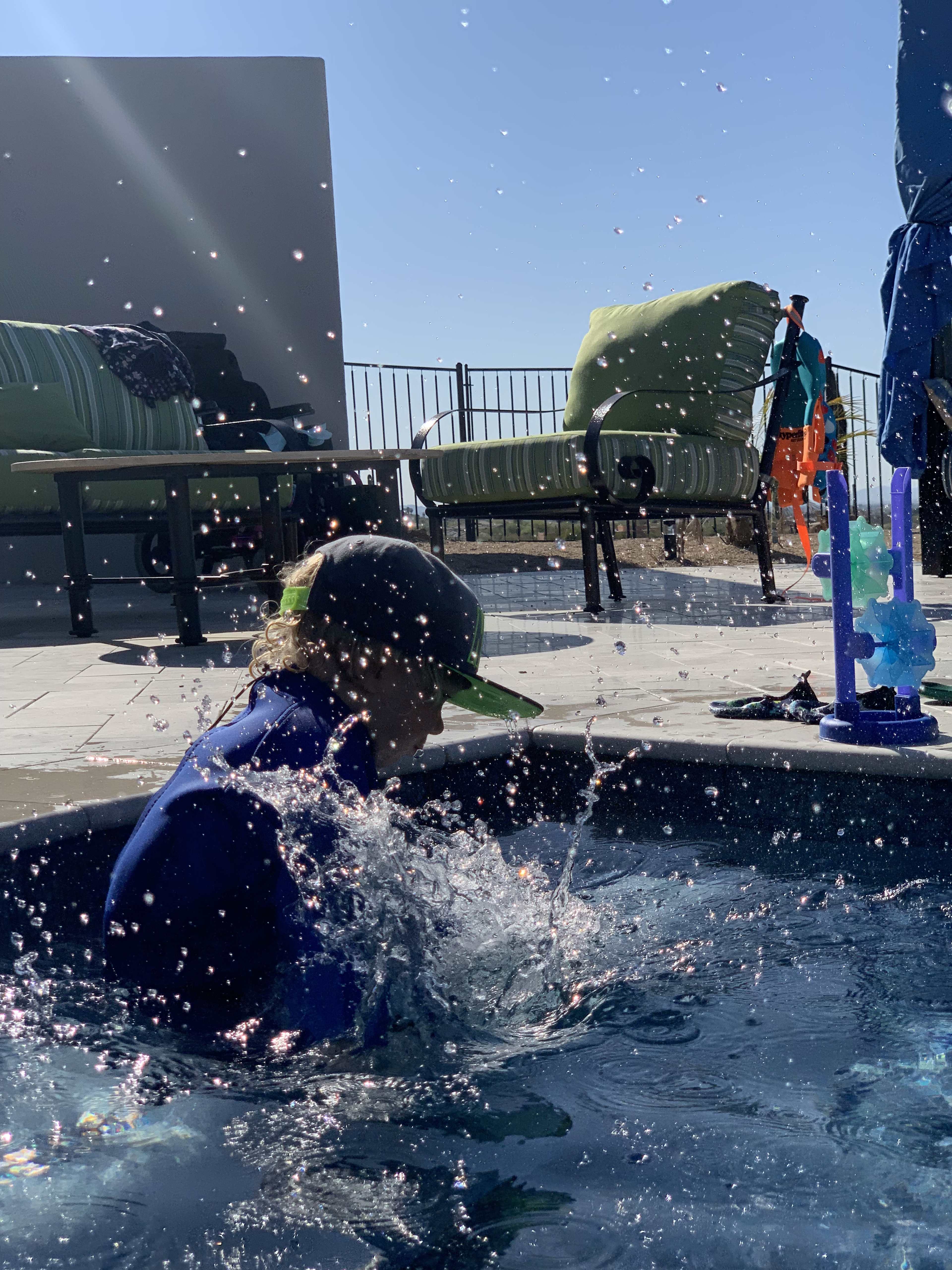A child splashes in a swimming pool, with patio furniture and toys visible in the background under a clear sky.