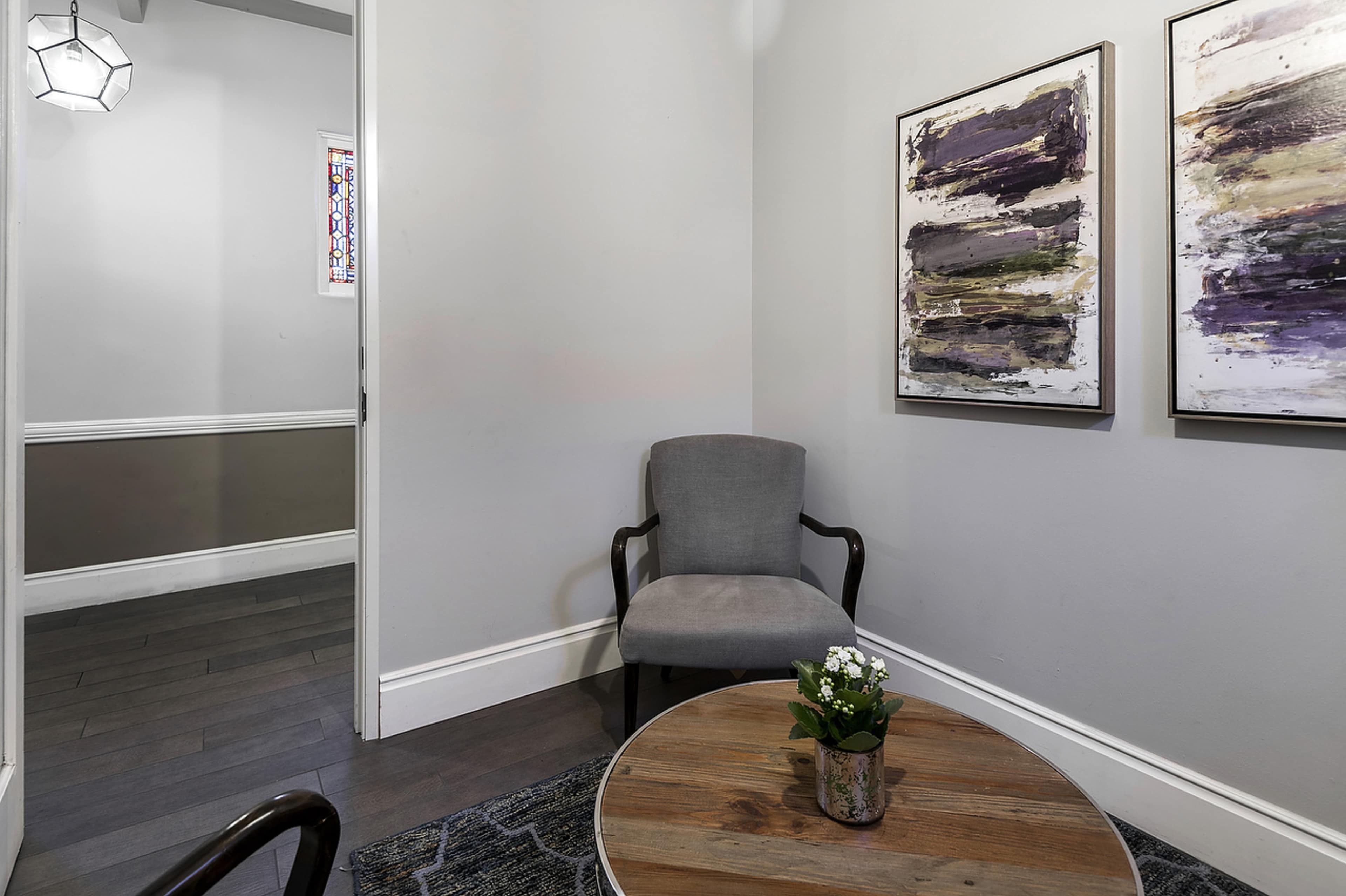 A small, modern waiting area features a gray chair, a round wooden table with a potted plant, and abstract artwork on the walls.
