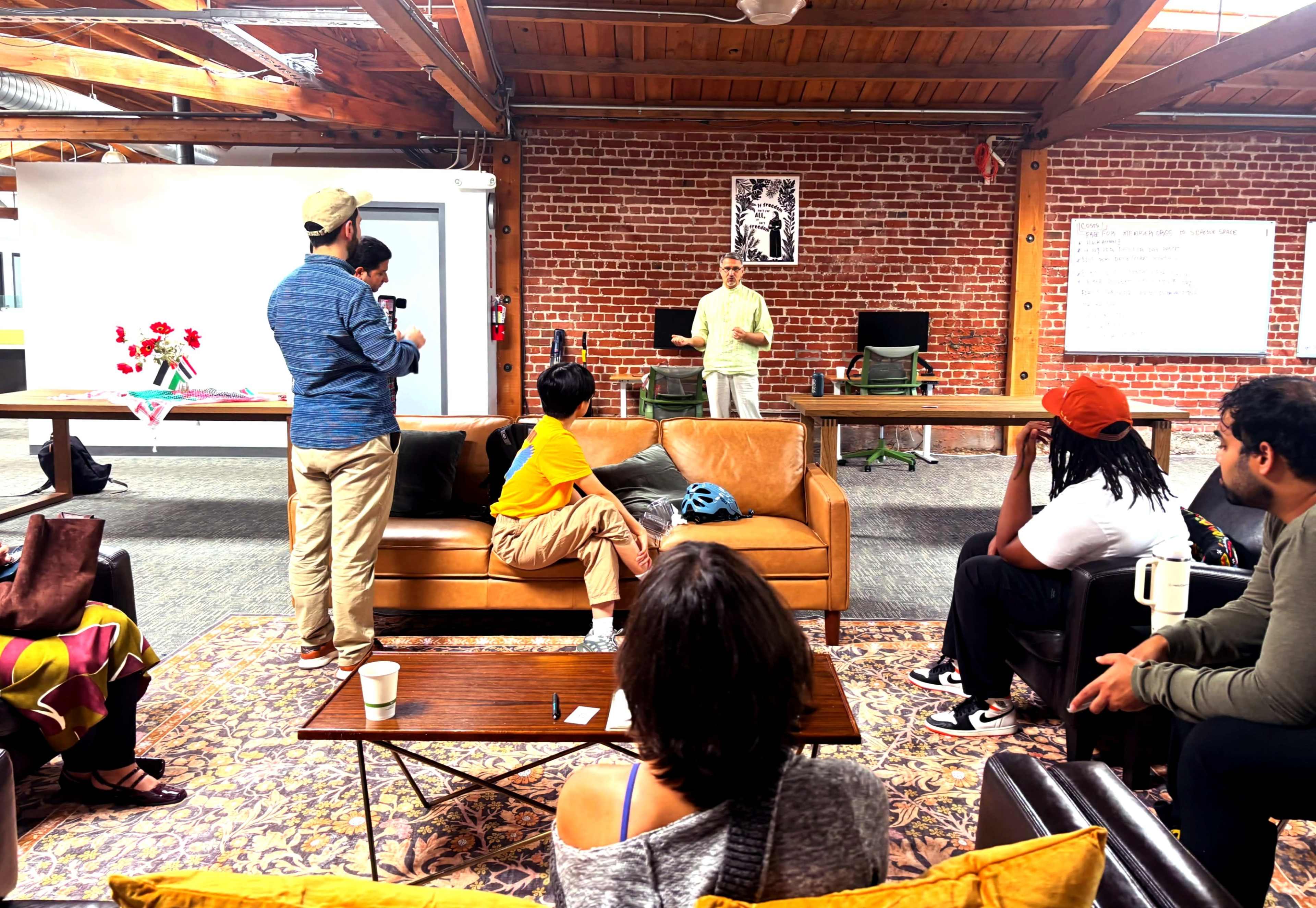 A group of people are gathered in a spacious room with brick walls and wooden beams, where one person is speaking while others listen attentively from various seating arrangements.
