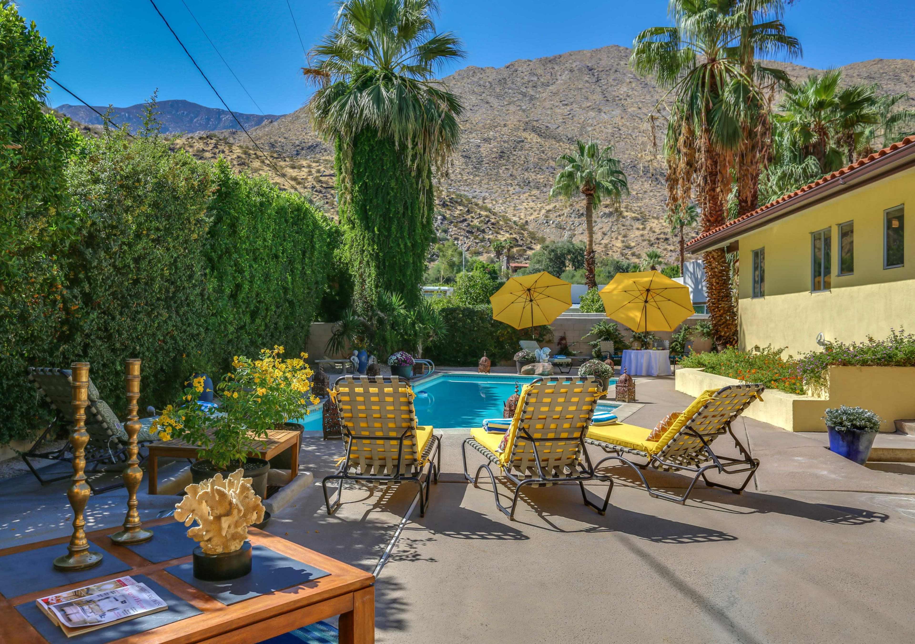 A swimming pool is surrounded by lounge chairs and decorative plants, with mountains in the background.