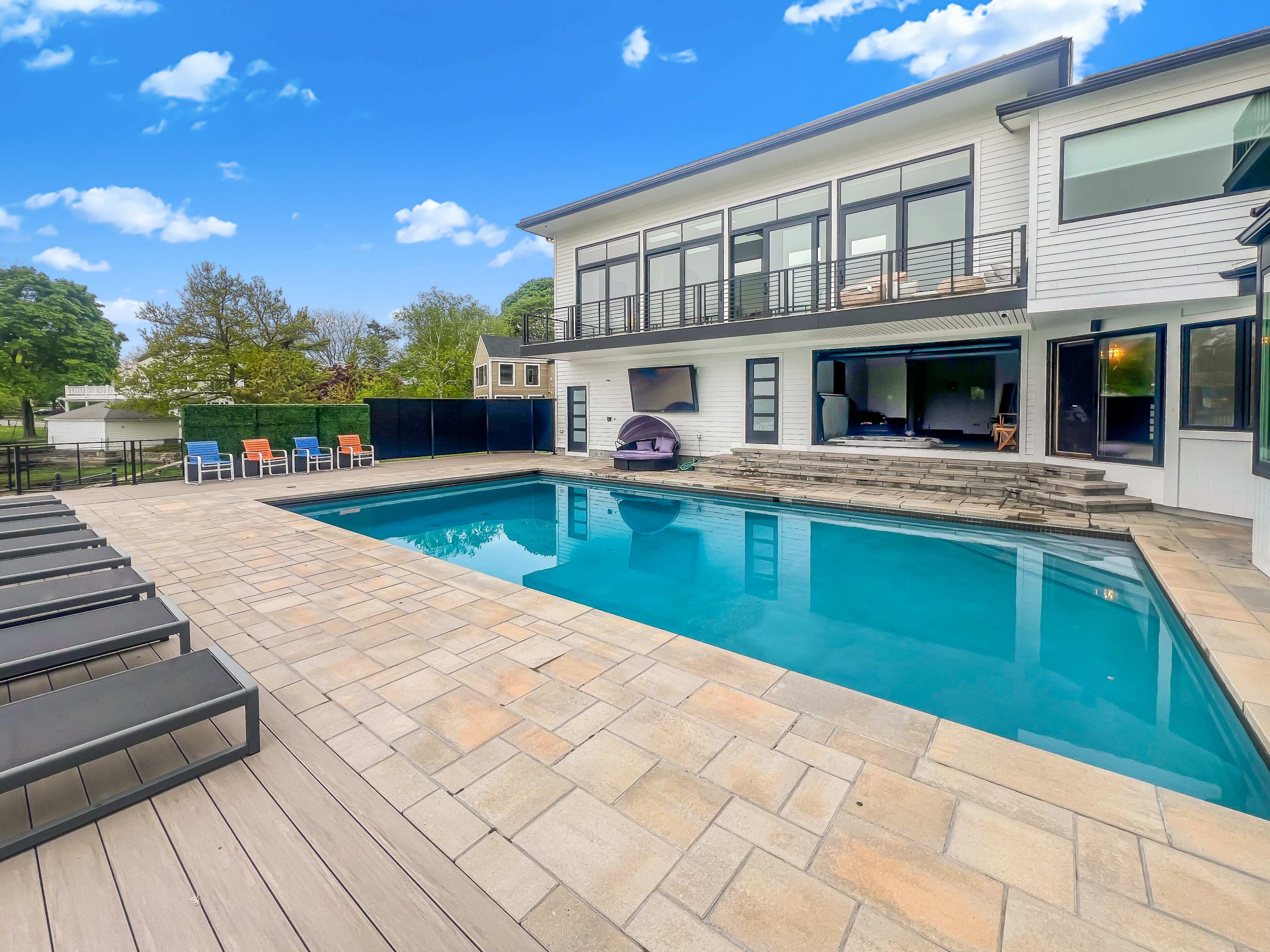 The image shows a modern house with a rectangular swimming pool surrounded by a stone patio and lounge chairs.
