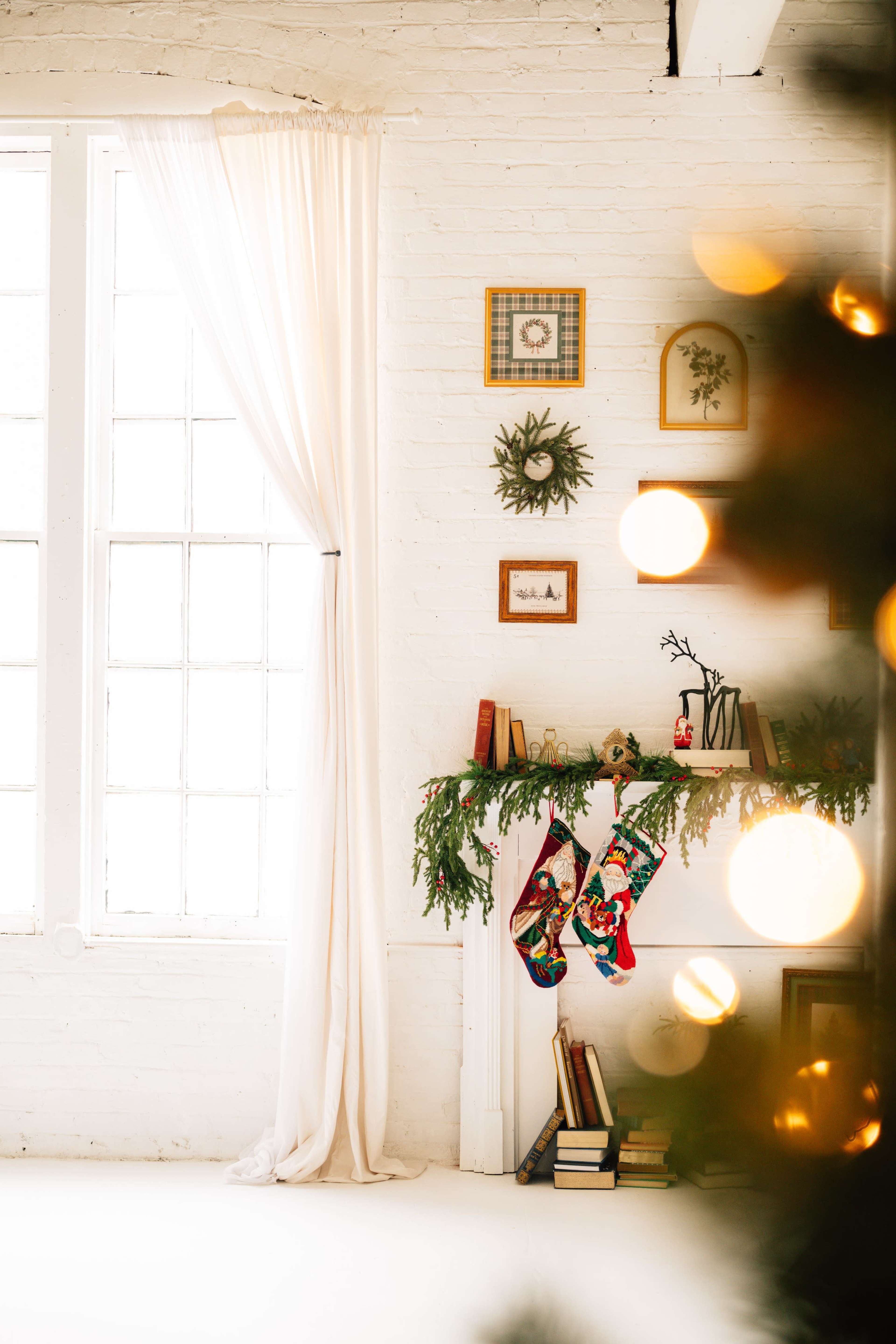A cozy room features a white brick wall with large windows, a decorated mantel with stockings, and a Christmas tree in the foreground.