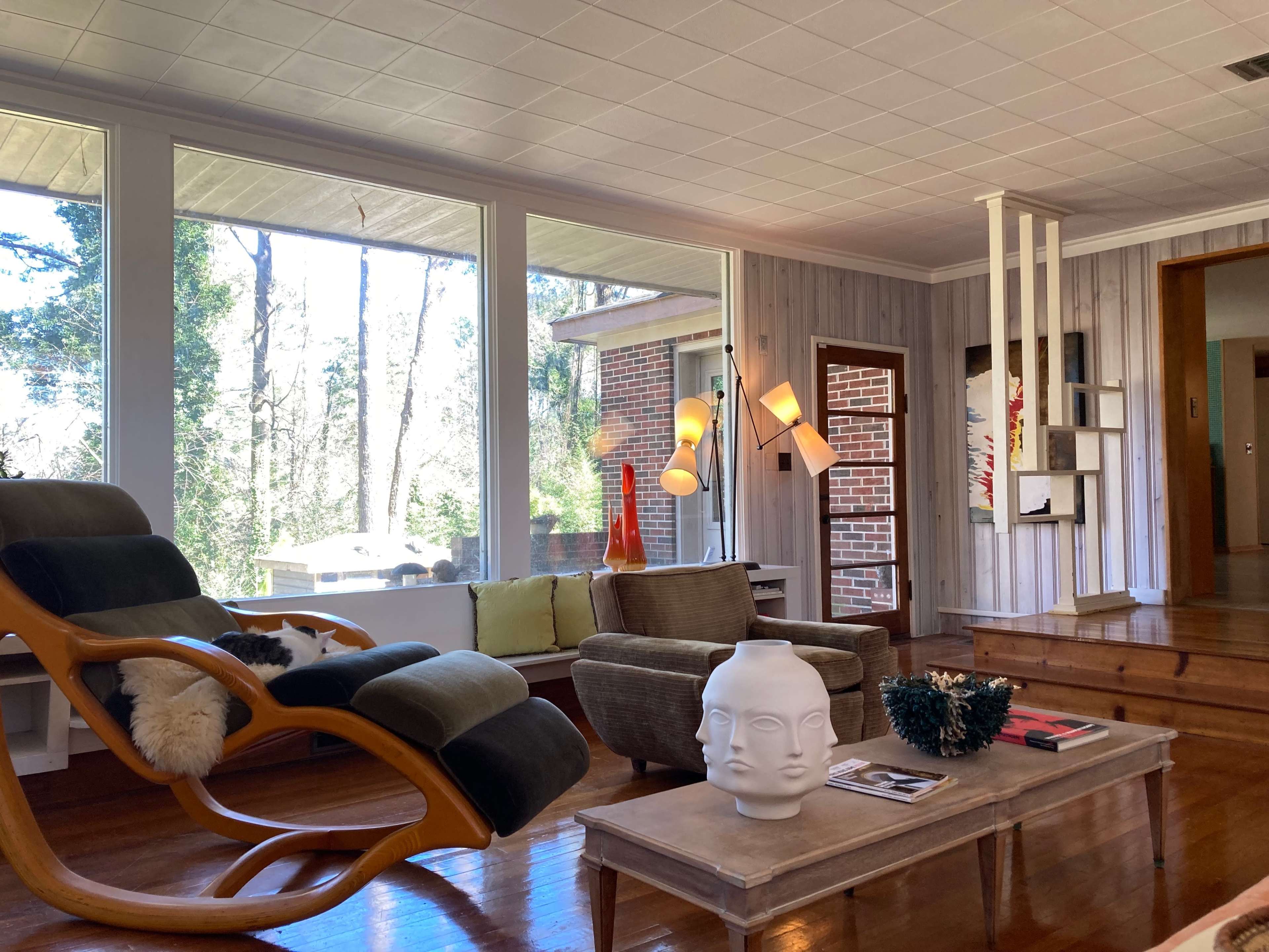 A modern living room features a wooden rocking chair, a green upholstered sofa, and a coffee table with a decorative head sculpture, all situated near large windows overlooking a wooded area.