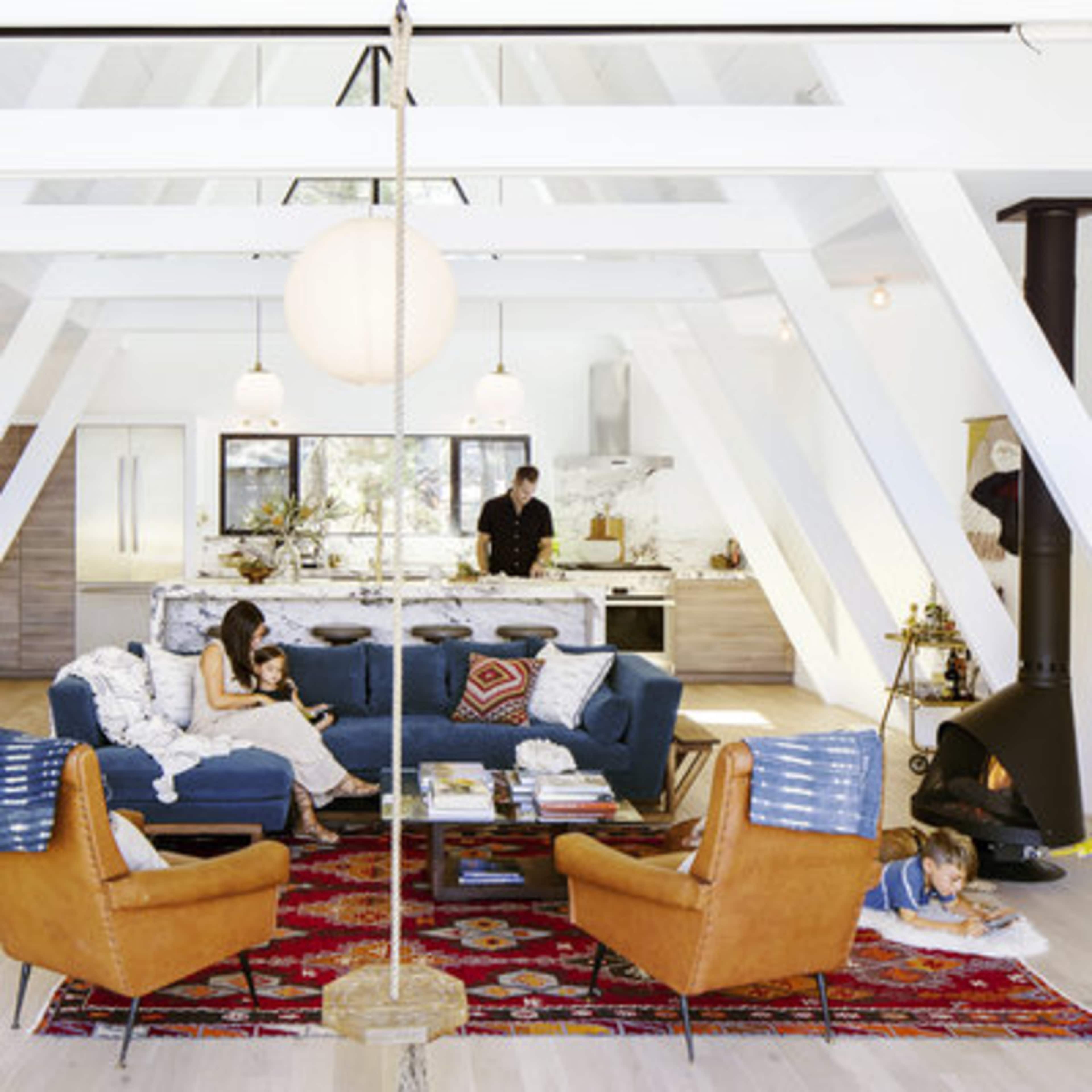 A brightly lit, modern living room features two orange armchairs and a blue sofa, with a kitchen visible in the background where a person is preparing food.