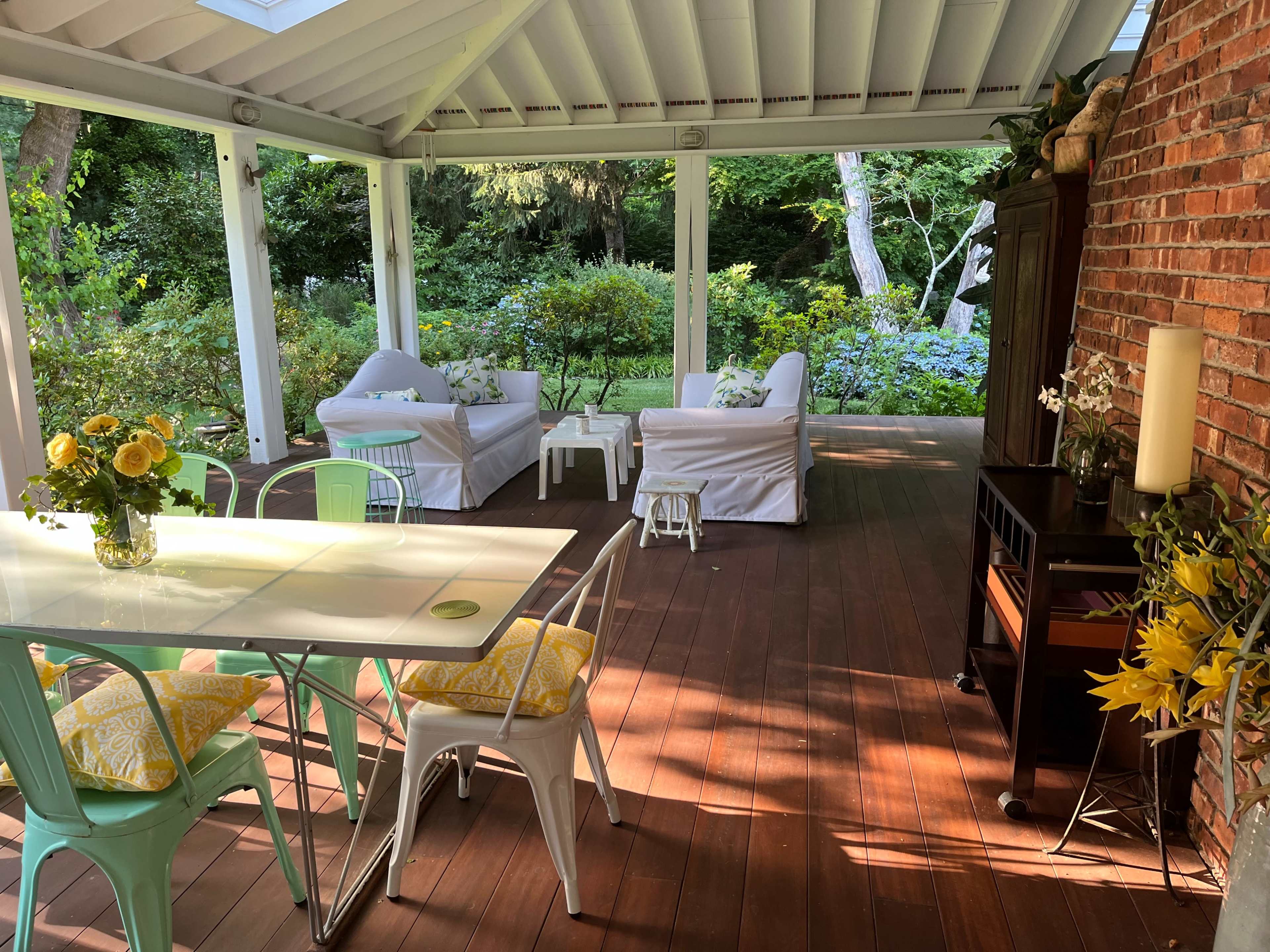 A covered wooden patio features a dining table with colorful chairs and a seating area with white couches surrounded by greenery.