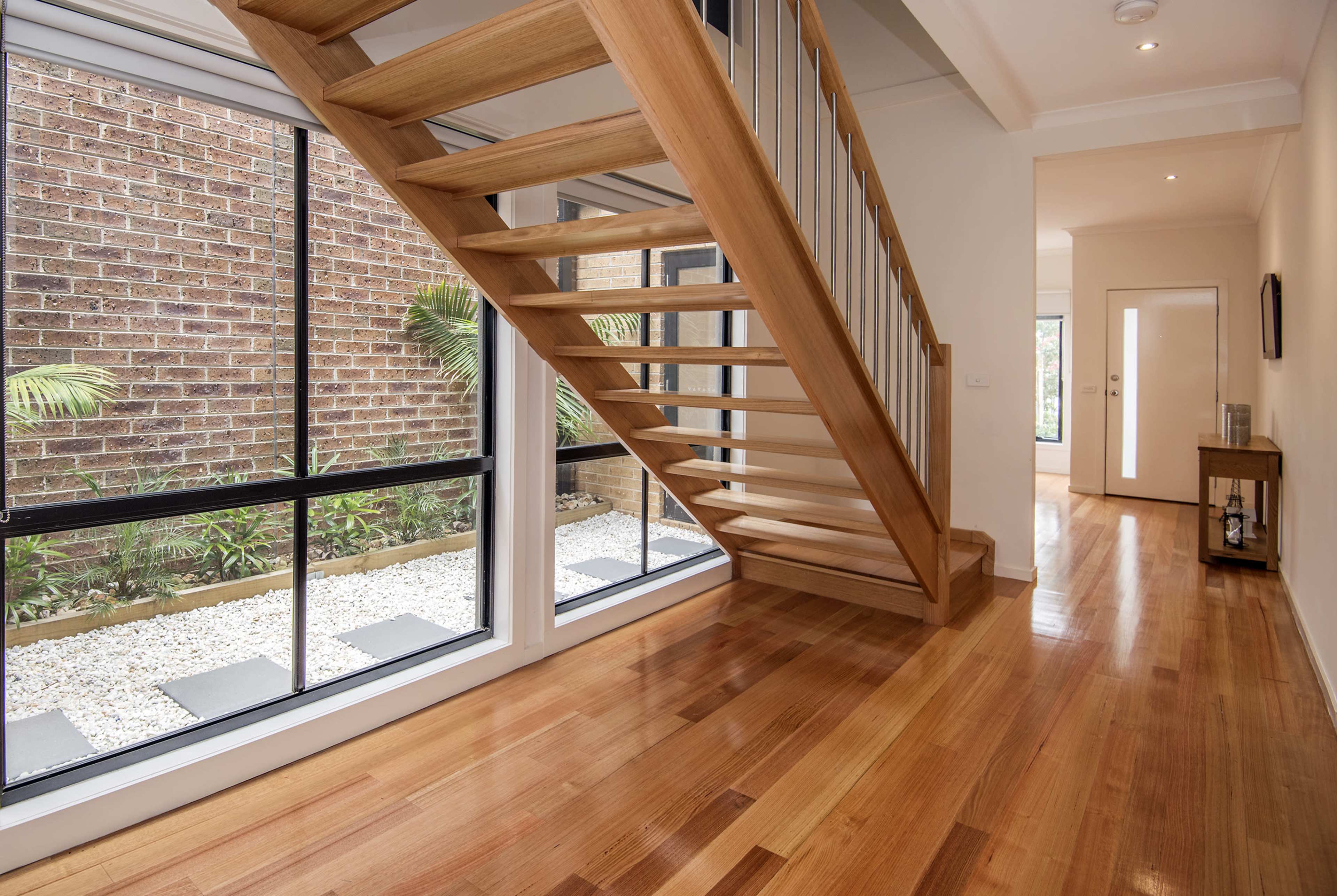 A modern interior hallway featuring a wooden staircase and large windows facing a gravel garden area.
