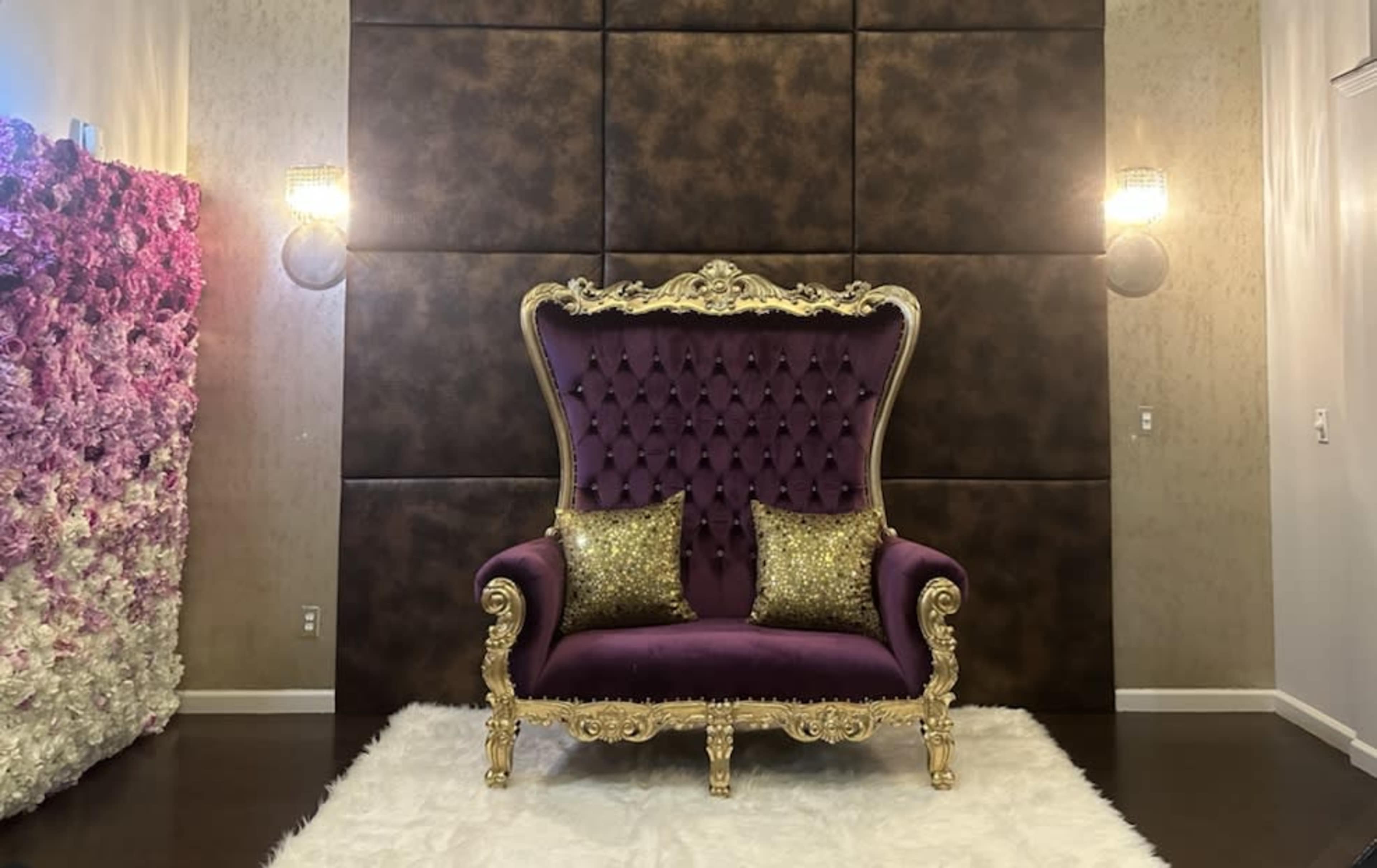 The image features a luxurious purple tufted sofa with ornate gold accents, positioned in front of a dark, textured wall and flanked by two decorative wall sconces.
