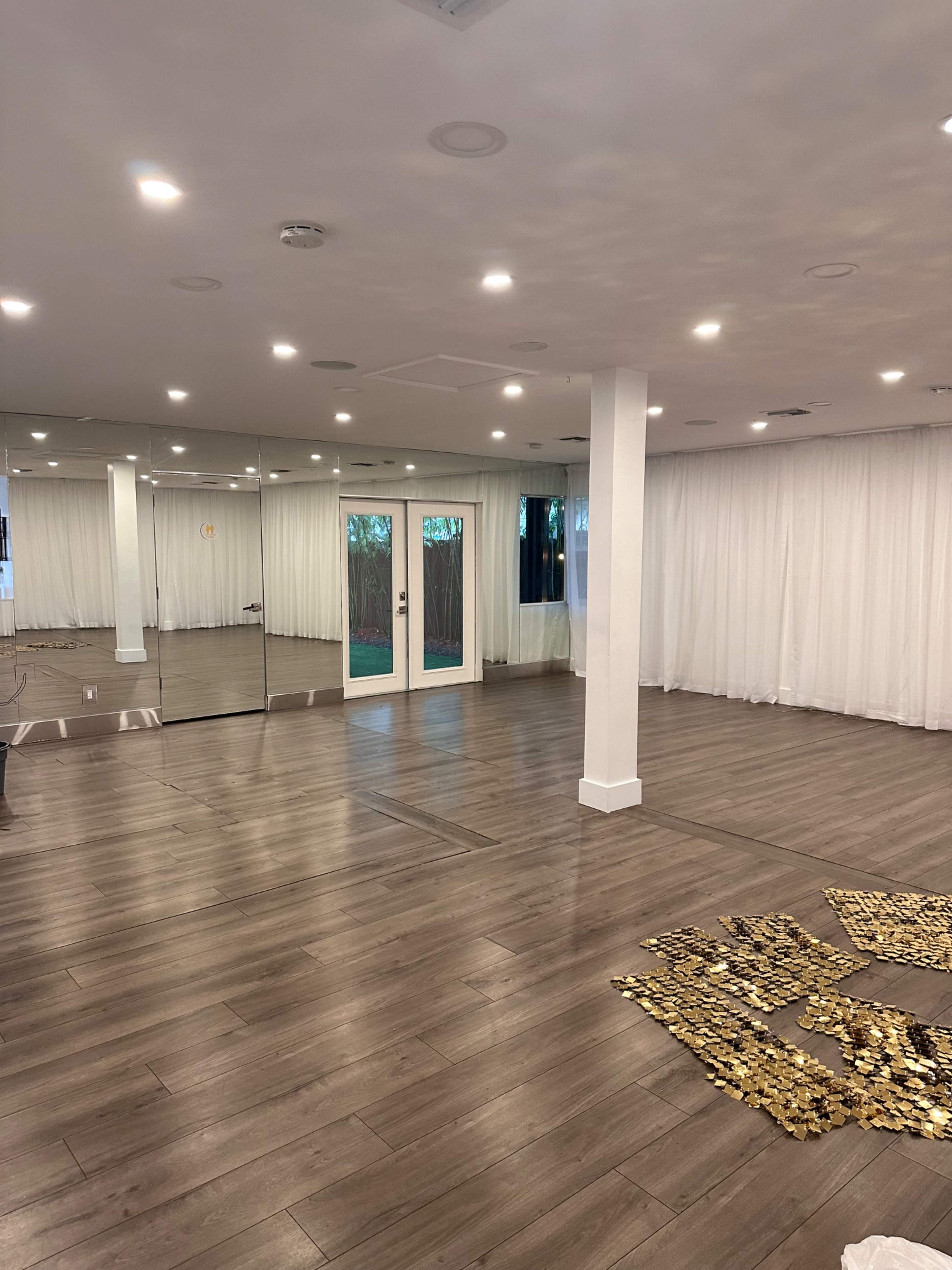 The room features a mirrored wall, large windows with sheer curtains, and a wooden floor, with a pattern arranged on the floor.