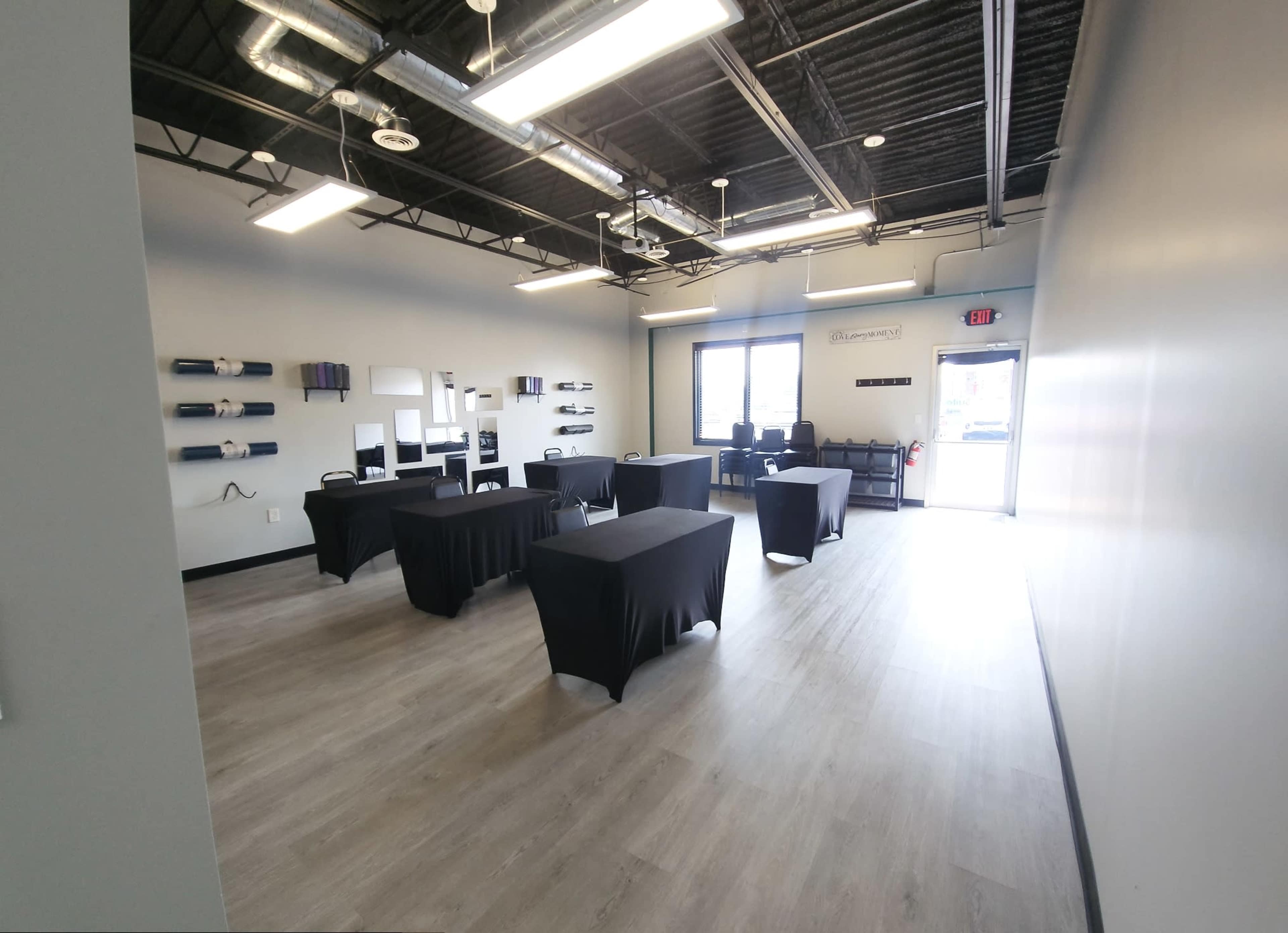 A spacious room with light gray walls, wooden flooring, and several black tables arranged in the center, leading to a door on the right side.