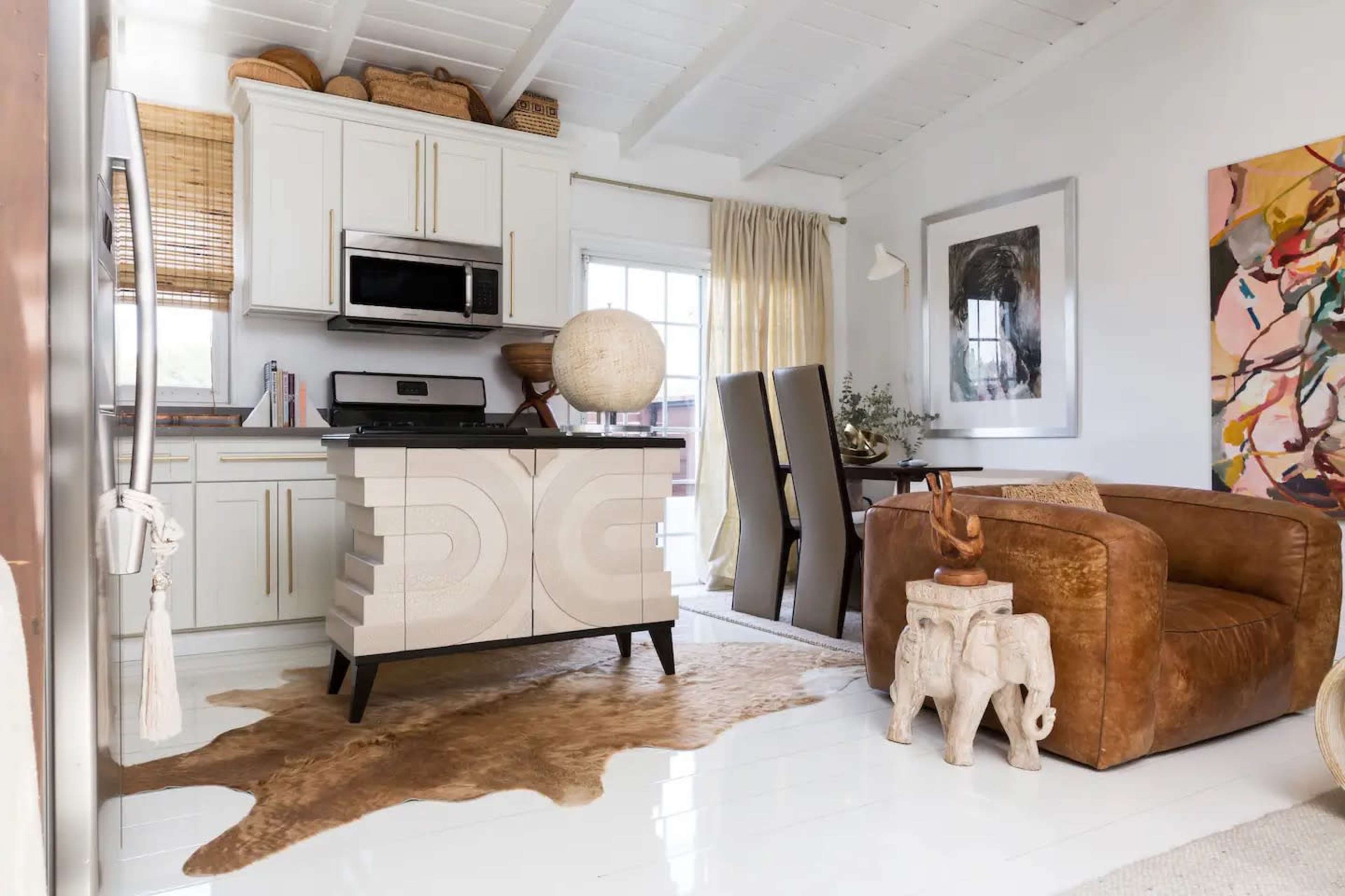 A modern kitchen features light cabinetry, a stainless steel microwave, and a round globe on the counter, with a dining area and a brown leather chair nearby.