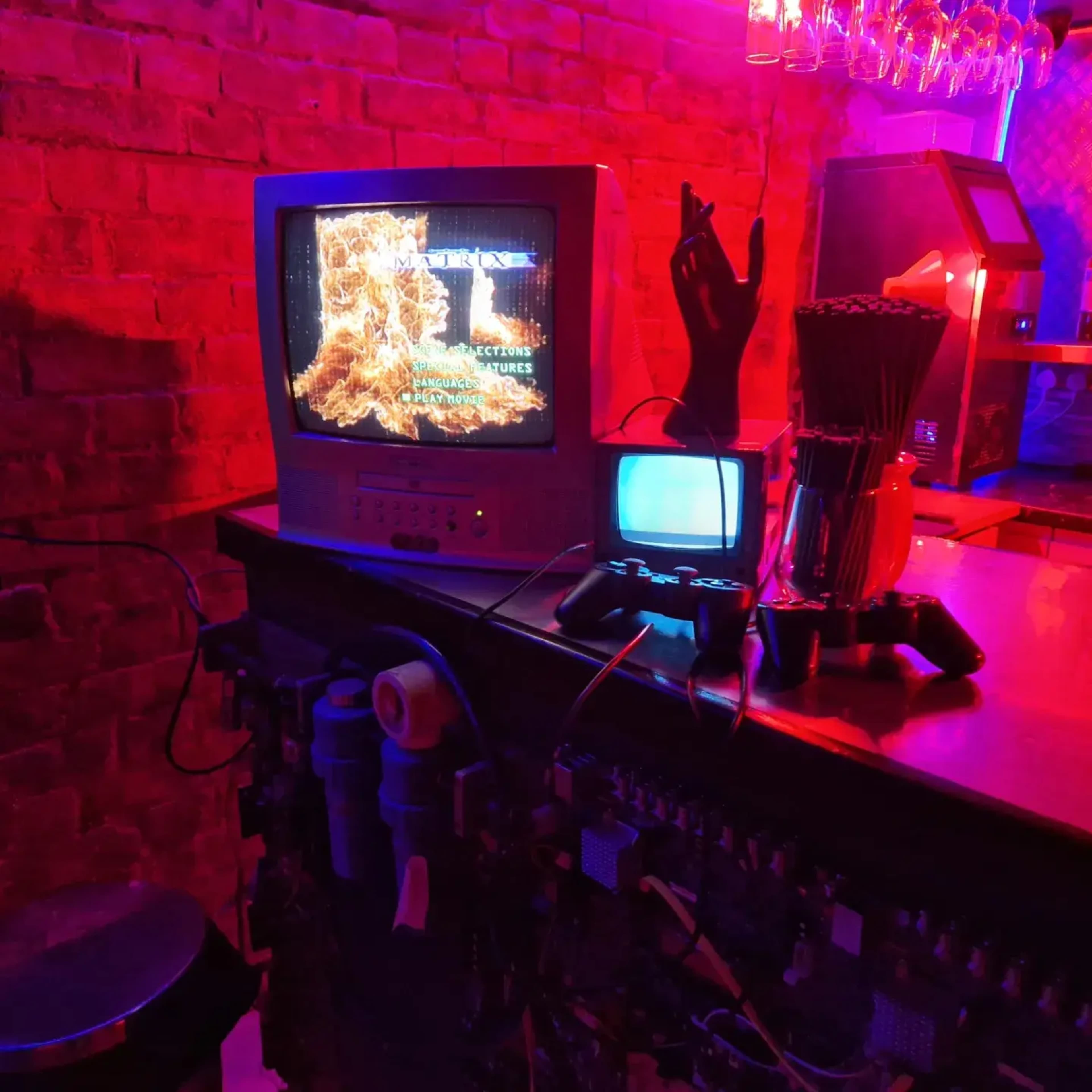 A vintage television displaying an image is placed on a bar counter alongside a smaller TV and a video game controller, with neon lighting and brick walls in the background.
