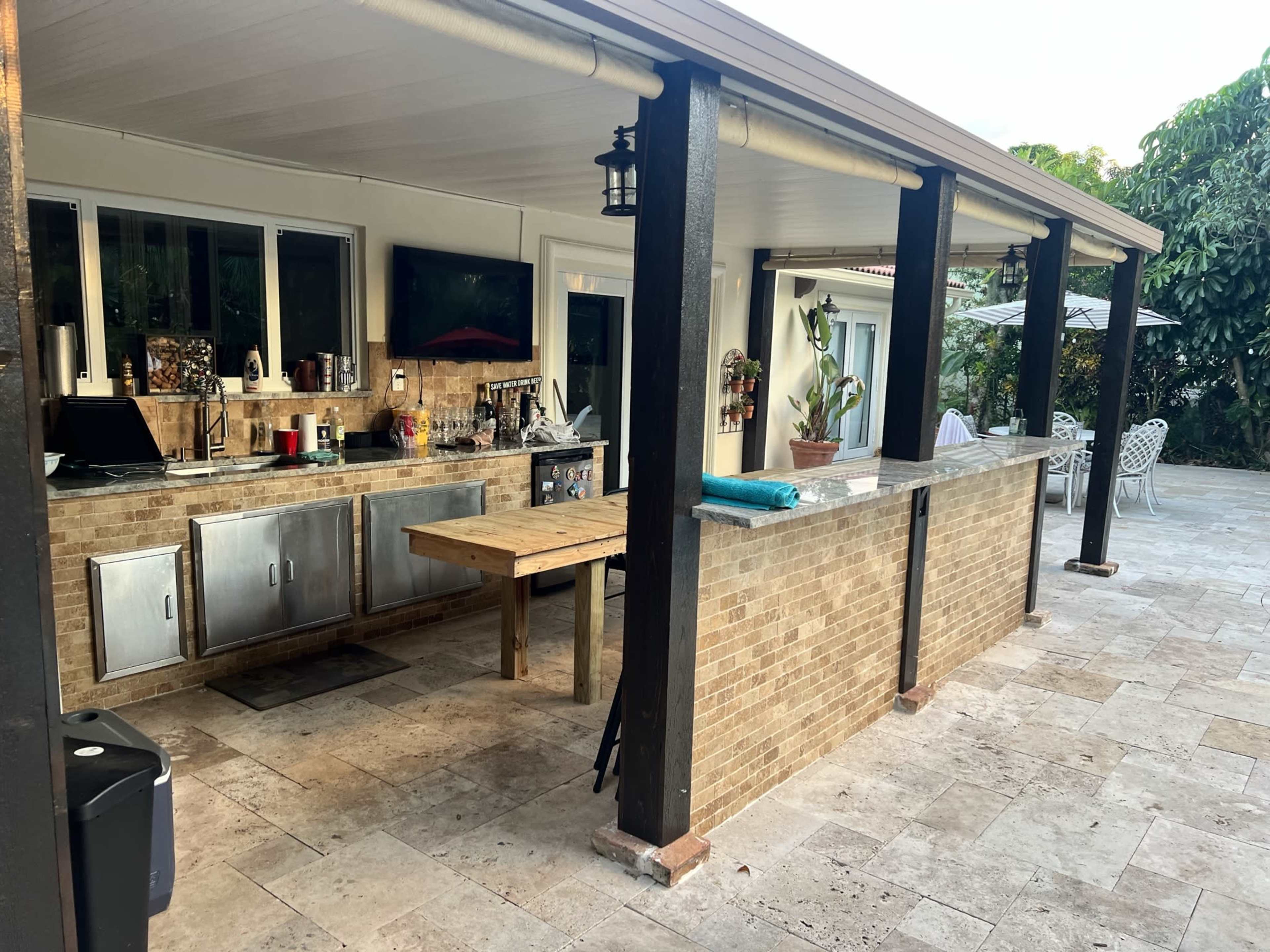 An open outdoor kitchen with a wooden table, stone countertops, and seating area surrounded by trees and patio furniture.
