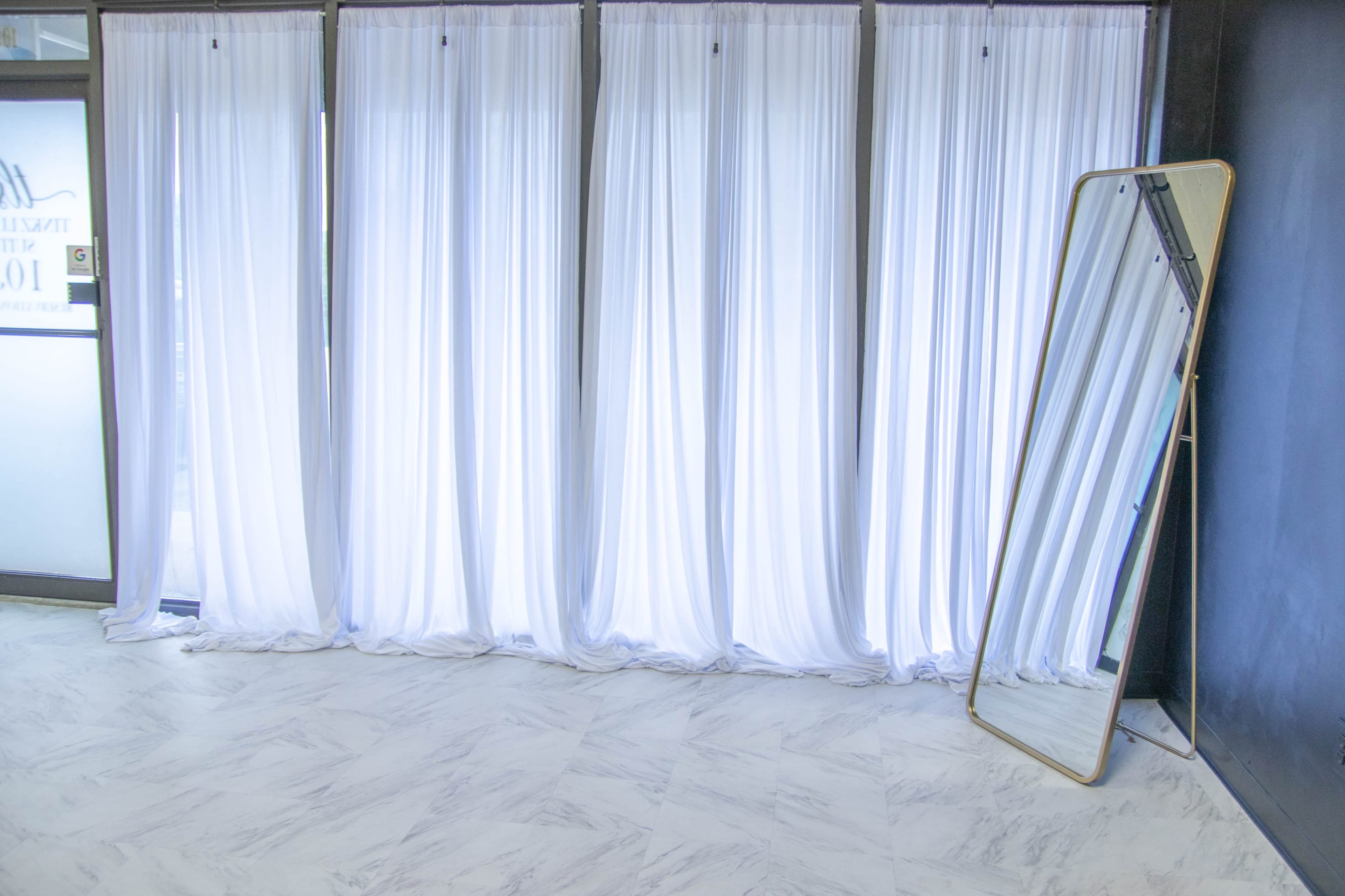 A spacious room with sheer white curtains covering large windows and a tall, framed mirror positioned on the floor.