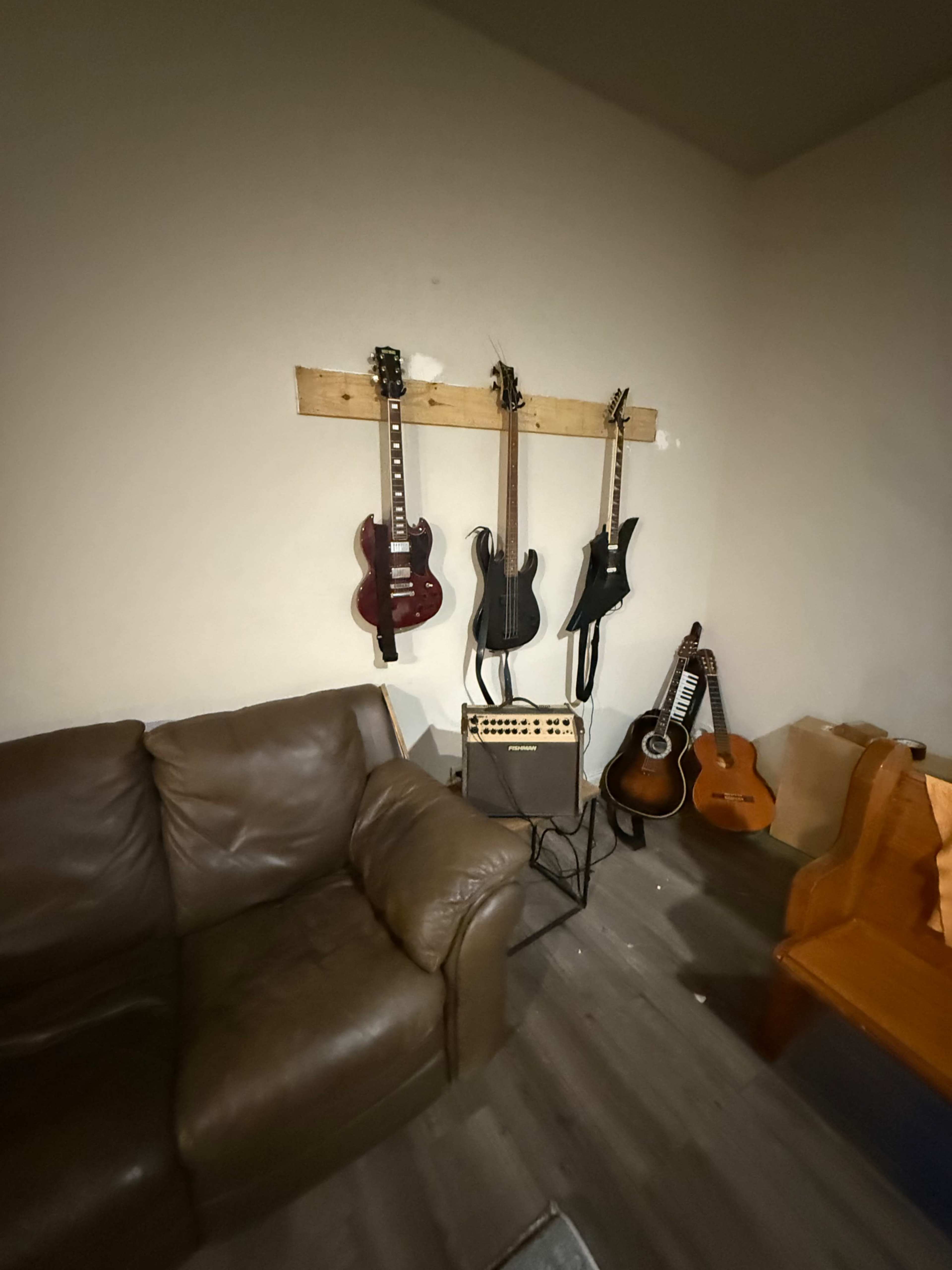 The image shows a cozy room with two brown sofas, a wooden wall-mounted rack holding three electric guitars, and an amplifier on the floor next to an acoustic guitar.
