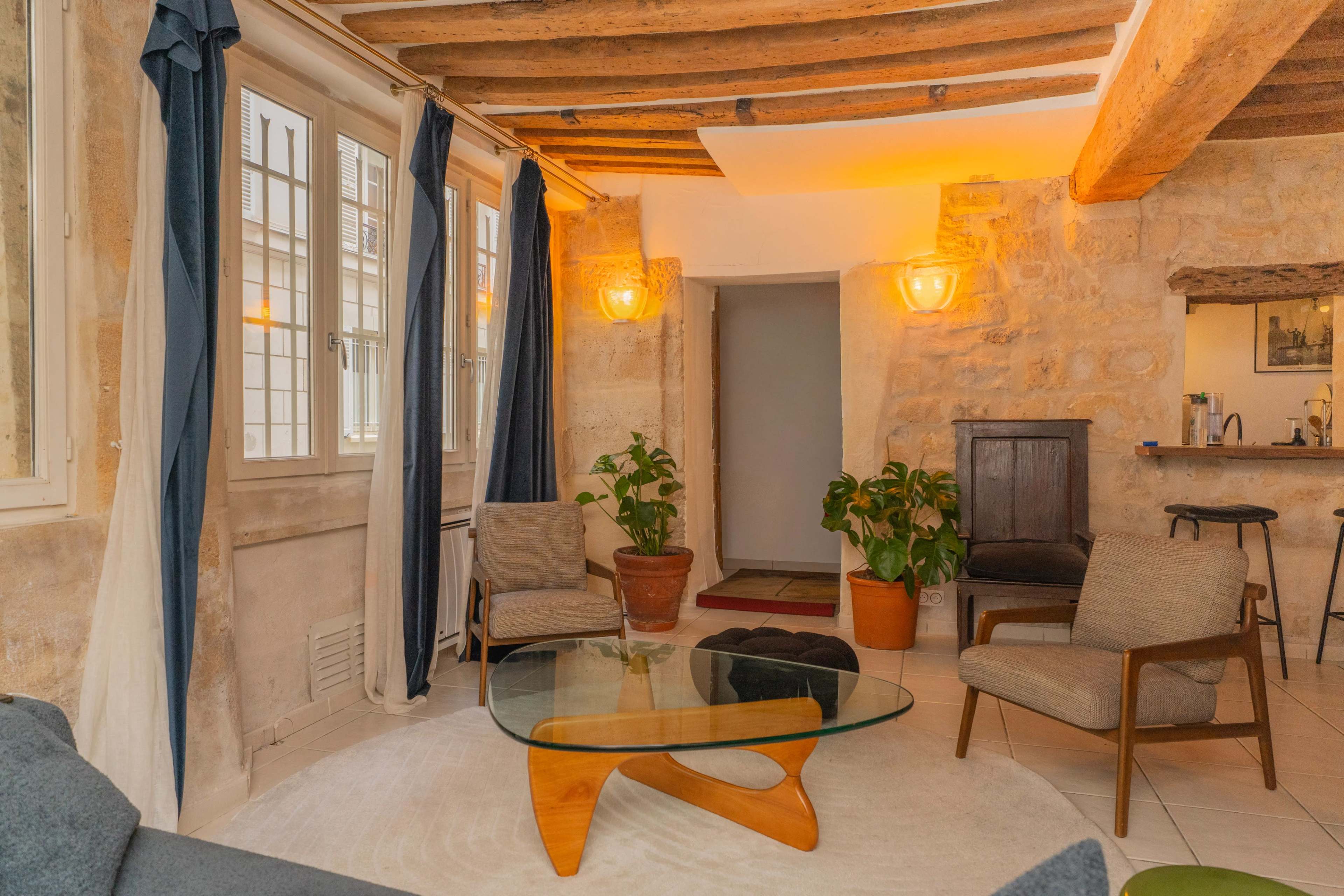 The image shows a cozy living room with two armchairs, a glass coffee table, and a large plant, surrounded by stone walls and wooden beams.