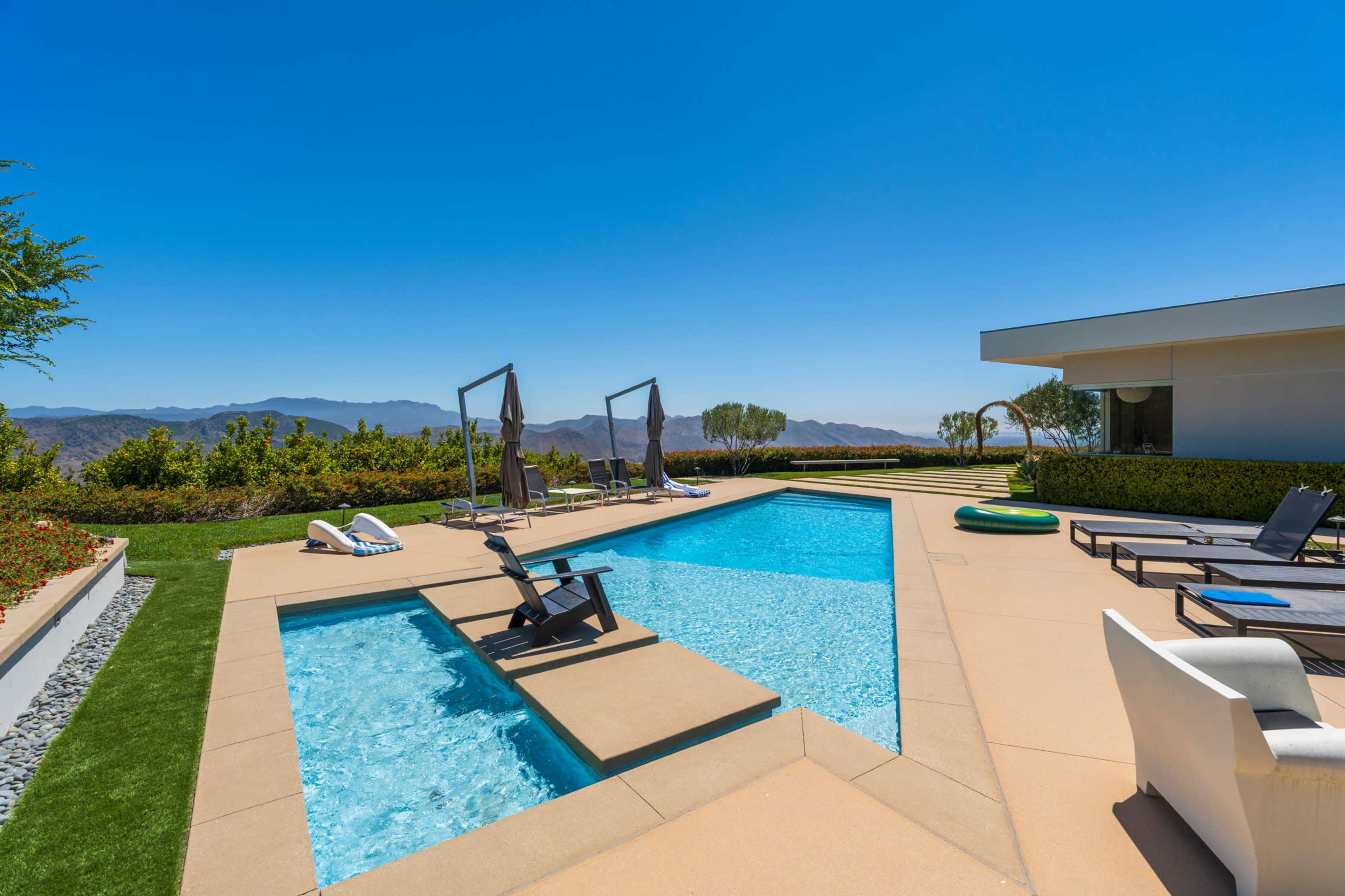 The image shows a modern backyard with a swimming pool, lounge chairs, and a landscaped area overlooking mountains and a clear blue sky.